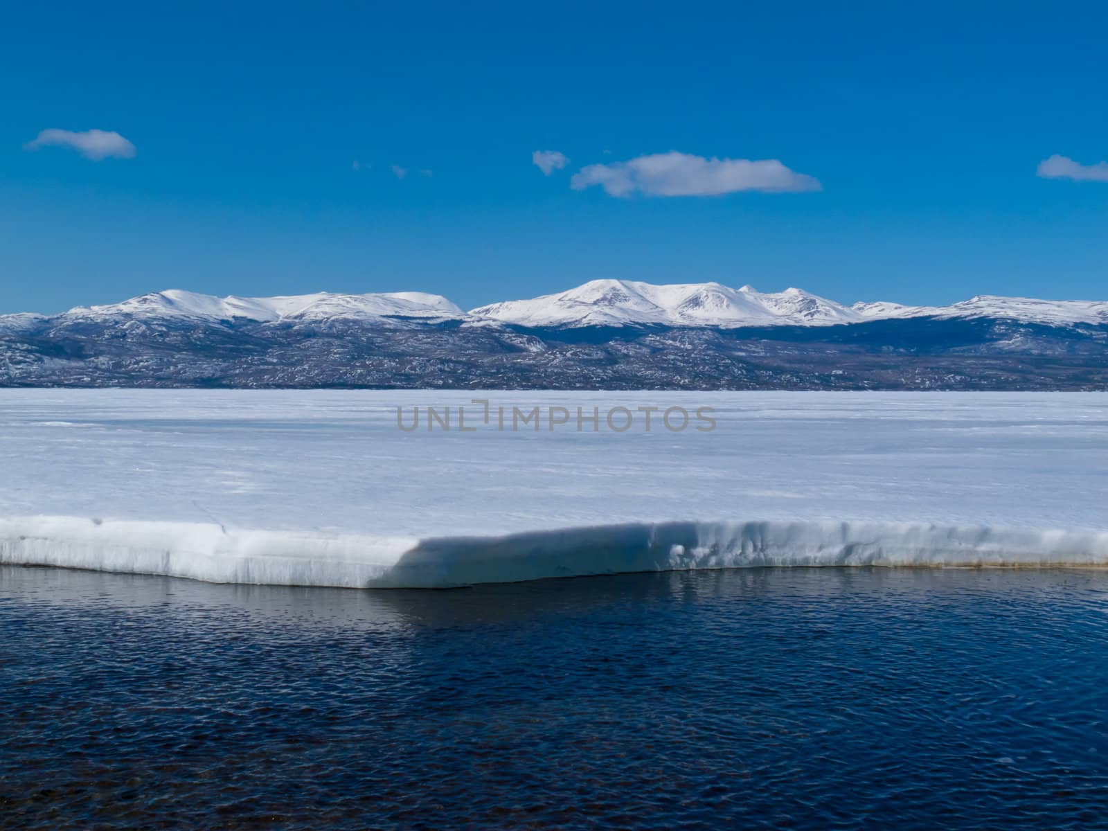 First open water at thawing Lake Laberge, Yukon Territory, Canada, and snow-covered mountains at distant shore of ice-covered frozen lake.