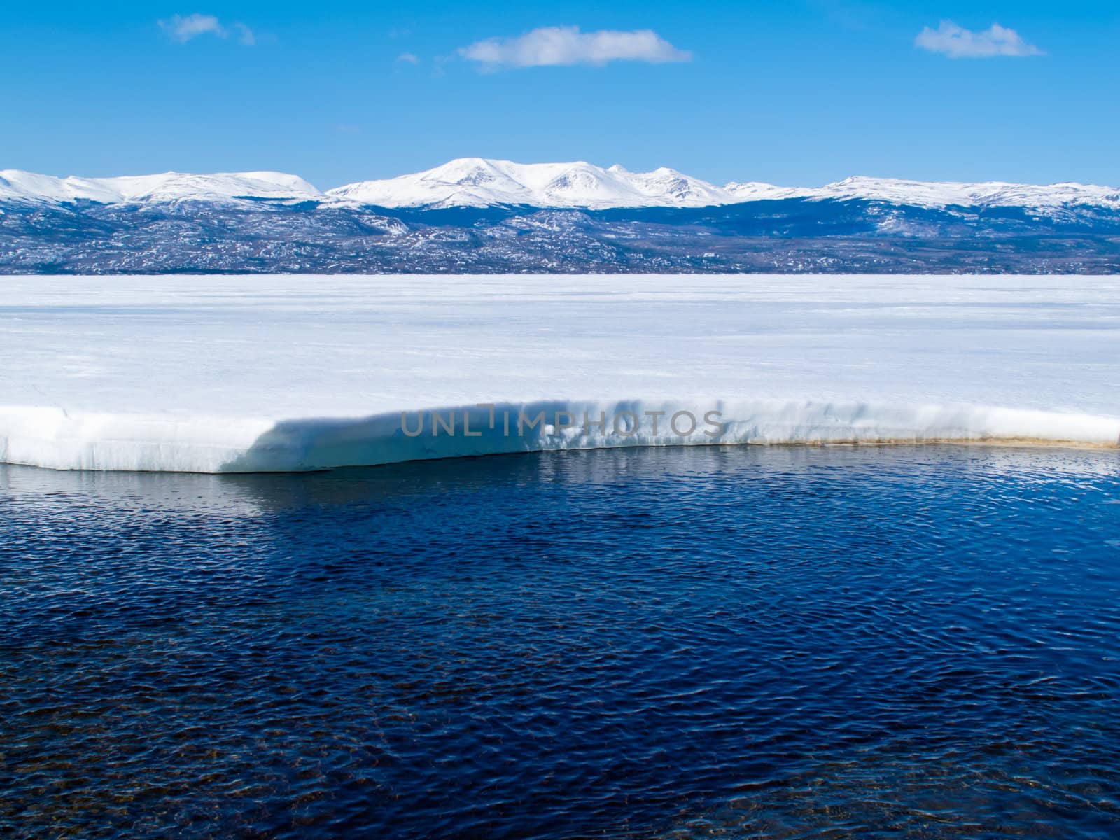 First open water at thawing Lake Laberge, Yukon Territory, Canada, and snow-covered mountains at distant shore of ice-covered frozen lake.