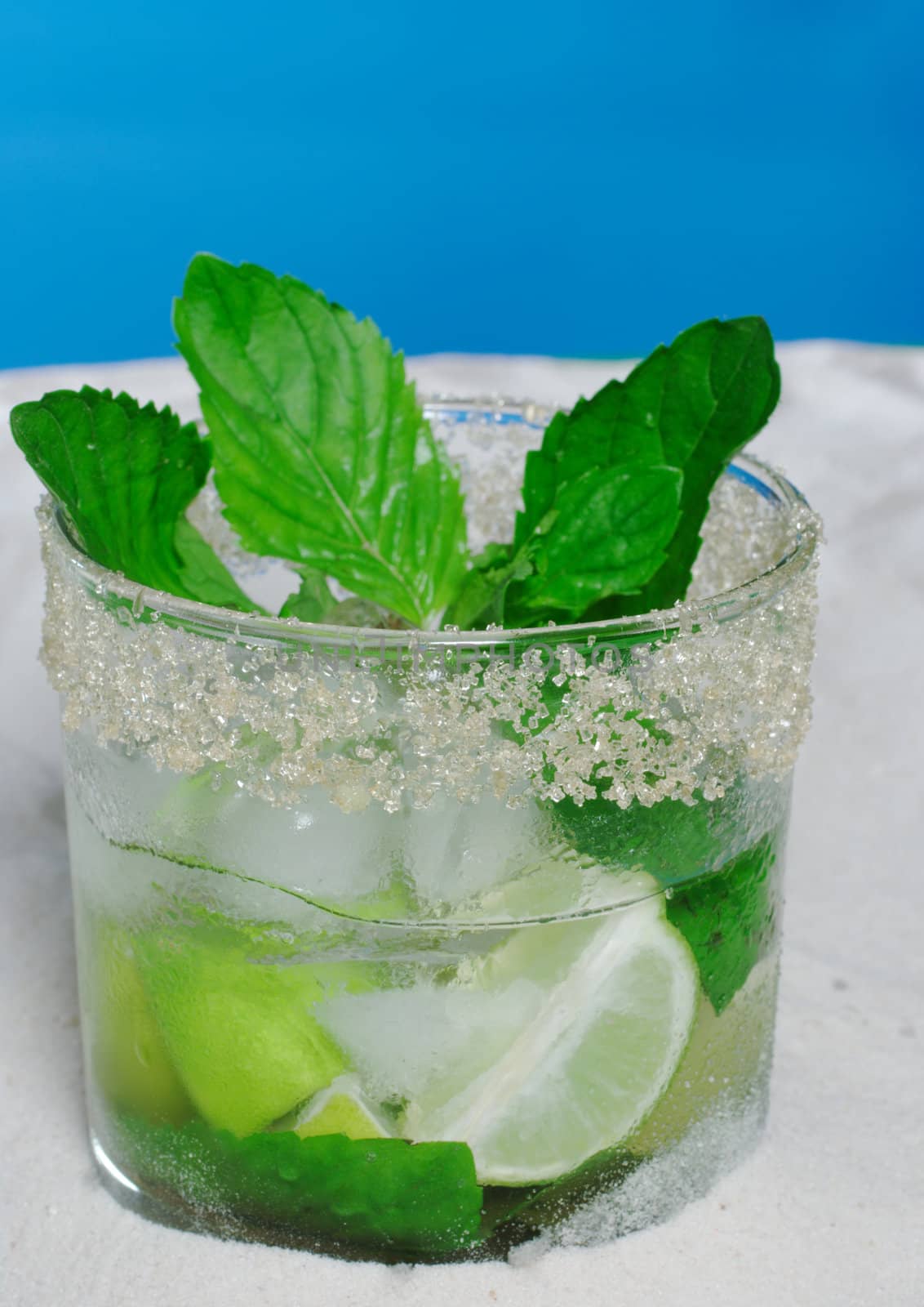 The Cocktail Mojito with Ice Cubes, Mint Leaves and Sugar Rim on Sand and with Blue Background 