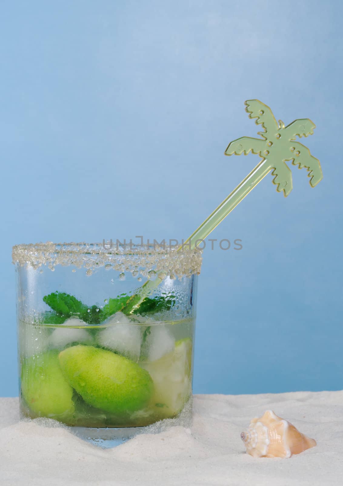 The Cocktail Mojito with Ice Cubes, Mint Leaves and Sugar Rim on Sand, with a palmtree stirring straw