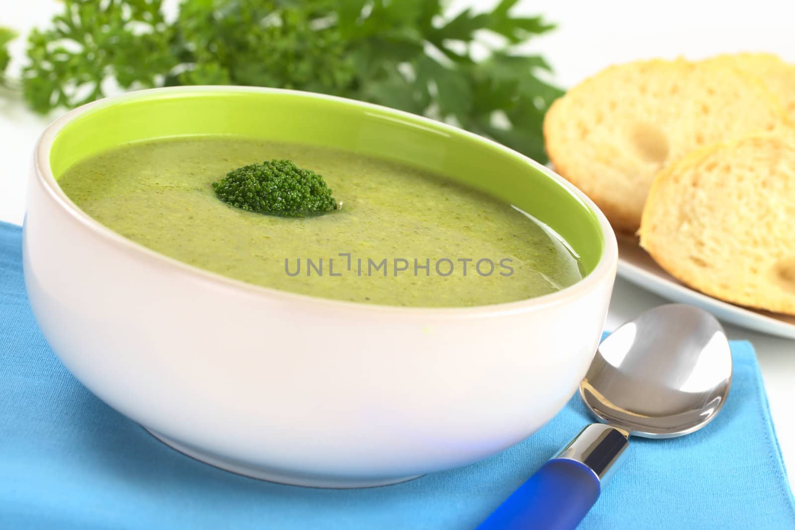 Cream of broccoli garnished with a broccoli floret on top with baguette slices and parsley leaves in the back (Selective Focus, Focus on the broccoli floret on the soup)