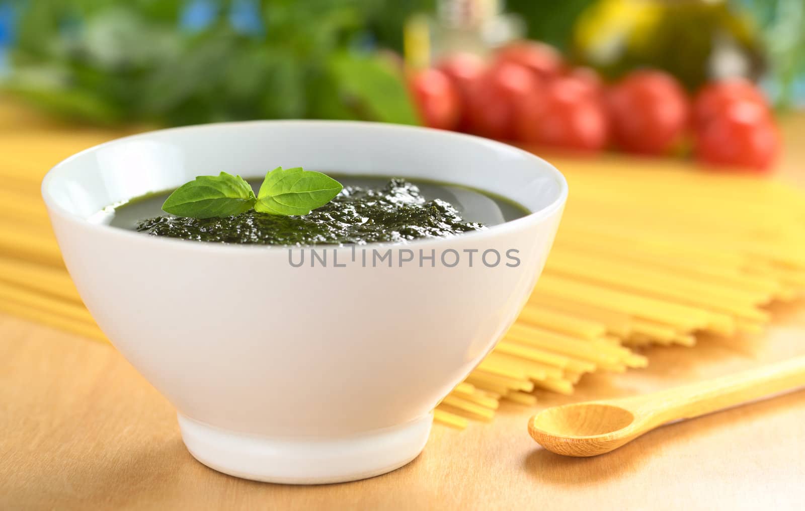 Fresh pesto made of basil, garlic and olive oil in a bowl and garnished with a basil leaf with raw pasta, cherry tomatoes, olive oil and basil leaves in the back (Selective Focus, Focus on the basil leaf in the bowl)