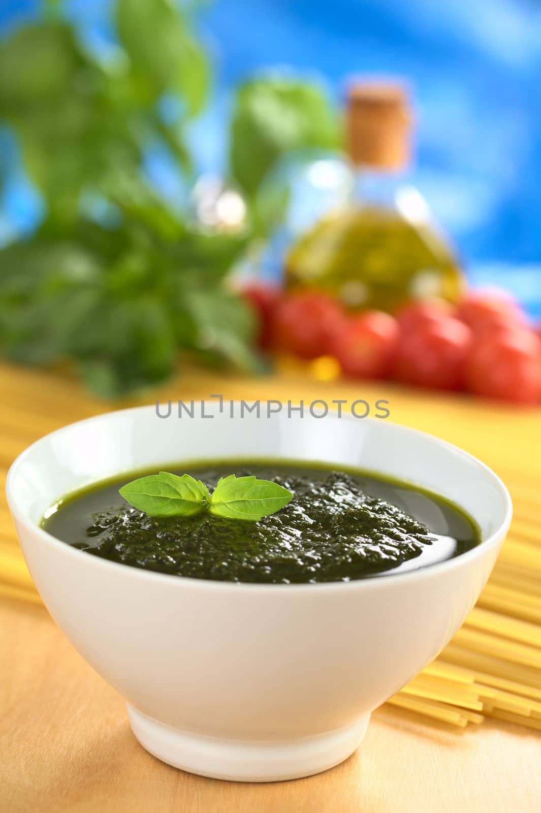 Fresh pesto made of basil, garlic and olive oil in a bowl and garnished with a basil leaf with raw pasta, cherry tomatoes, olive oil and basil leaves in the back (Selective Focus, Focus on the basil leaf in the bowl)