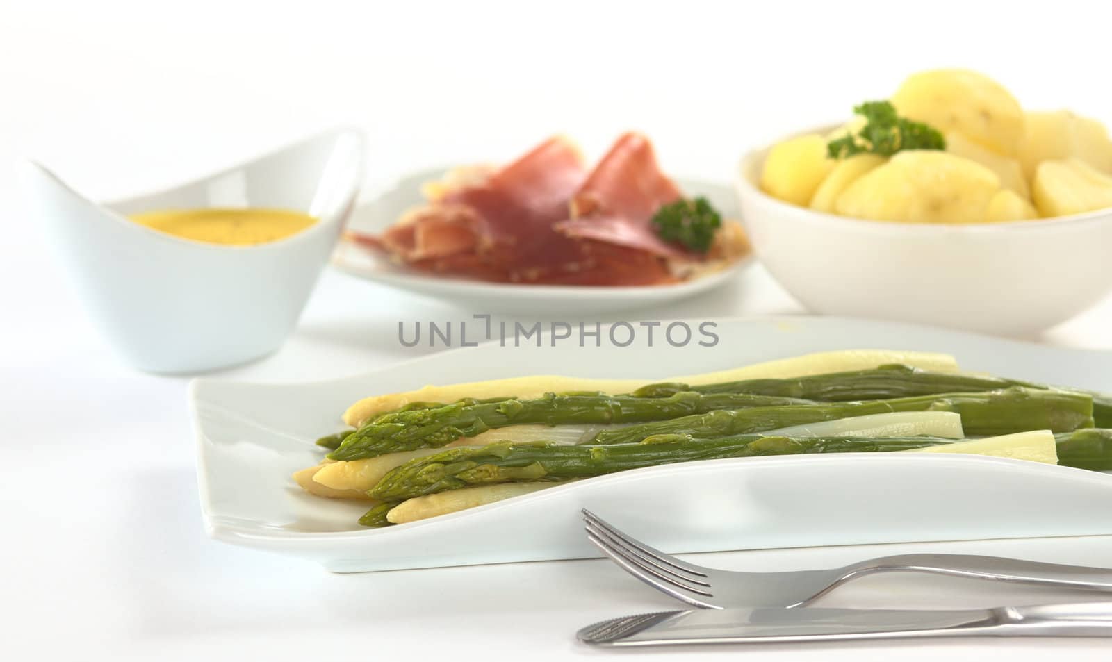 Cooked white and green asparagus on long plate with fork and knife in front and potatoes, ham and Hollandaise sauce in the background (Selective Focus, Focus on the front of the asparagus)
