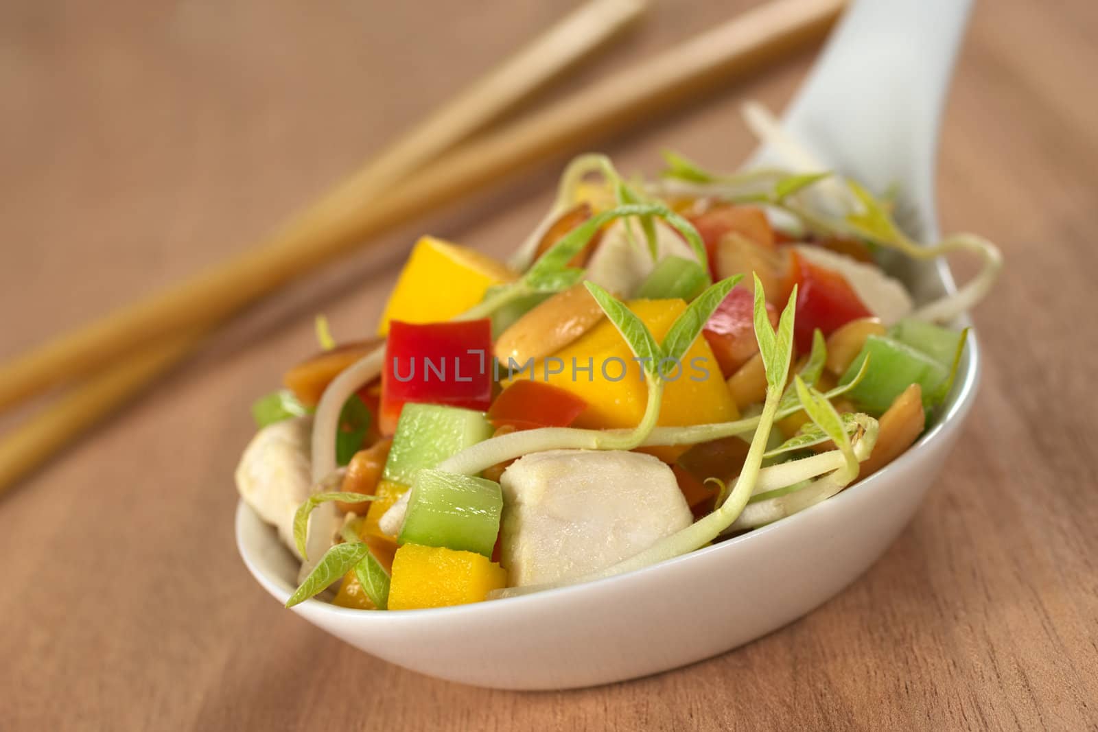 Fresh Asian salad with chicken, mango, cucumber, bean sprouts, red bell pepper and peanuts on white ceramic spoon with wooden chopsticks in the back (Selective Focus, Focus on the front of the food)
