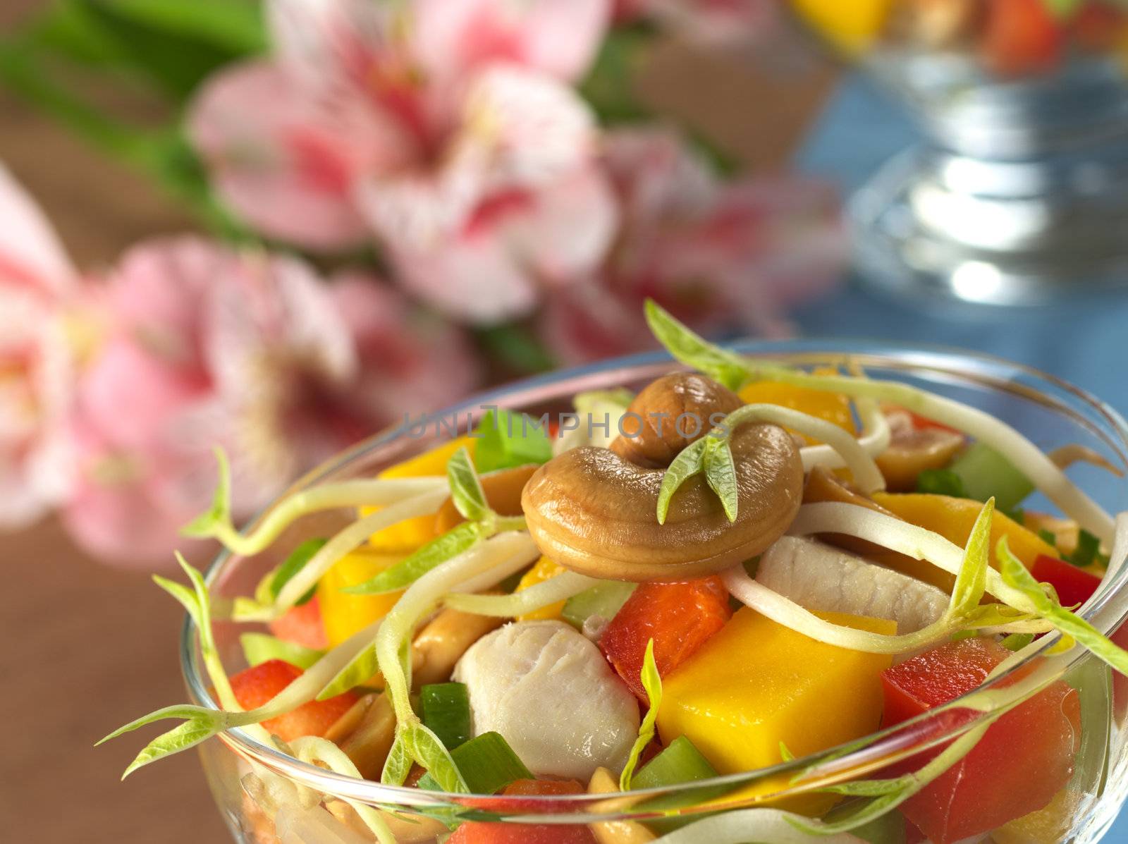 Fresh Asian salad with chicken, mango, cucumber, bean sprouts, red bell pepper and peanuts in glass bowl with inca lily in the back (Selective Focus, Focus on the cashew nut and the bean sprout on the top)