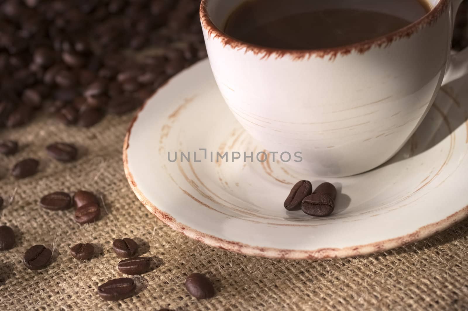 Coffee beans on the saucer of a coffee cup surrounded by many coffee beans on fabric called Jute (Very Shallow Depth of Field, Focus on the front coffee bean on the saucer and some on the fabric)