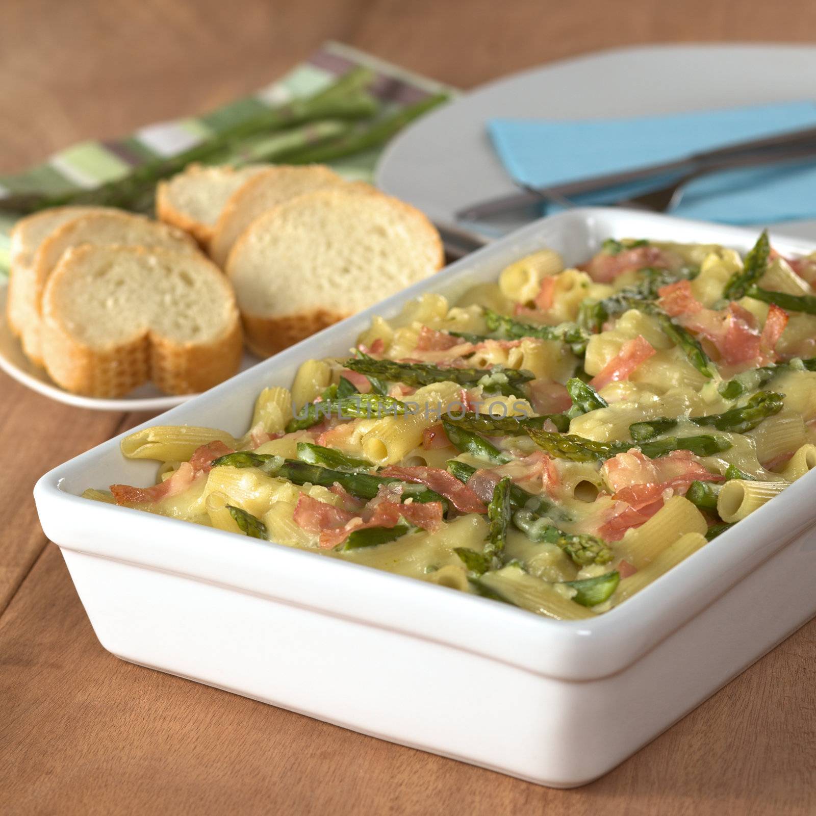 Casserole of green asparagus, ham and macaroni with baguette slices and plates in the back (Selective Focus, Focus in the middle of the dish)