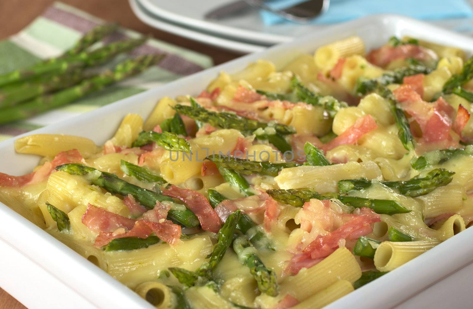Casserole of green asparagus, ham and macaroni with raw green asparagus and plates in the back (Selective Focus, Focus one third into the dish)