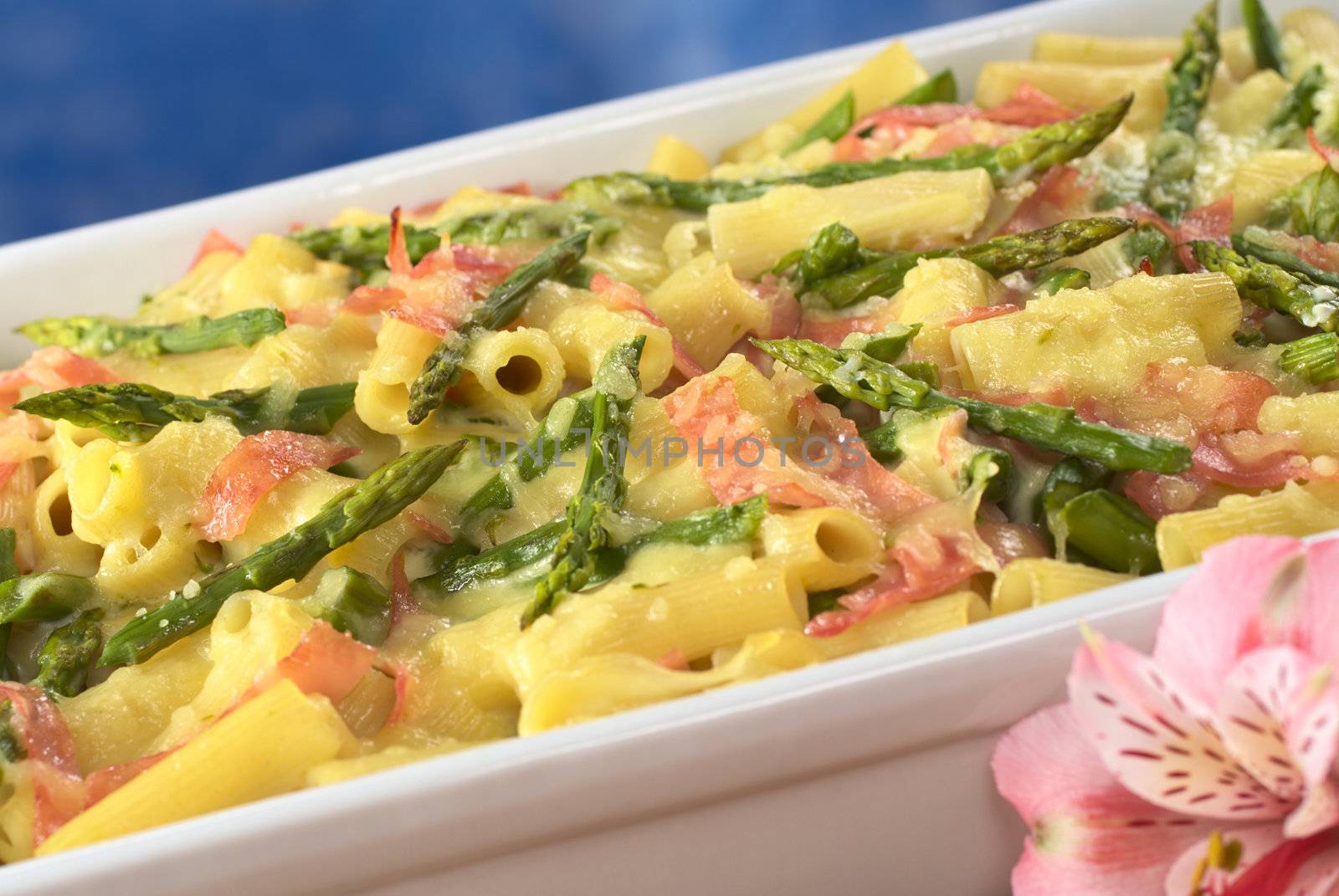 Green asparagus-ham-macaroni casserole with grated cheese on top and a pink Inca Lily in front (Selective Focus, Focus one third into the dish)