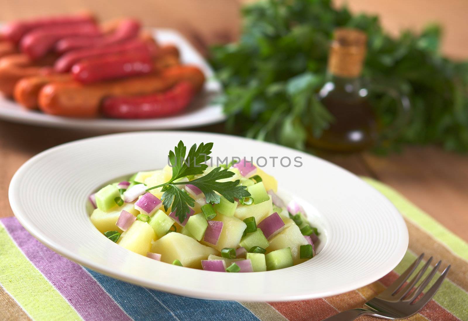 Fresh potato salad with cucumber, red and green onion made with an oil dressing with barbecued sausages, oil and parsley in the back (Selective Focus, Focus on the parsley leaf on the salad and its surroundings)
