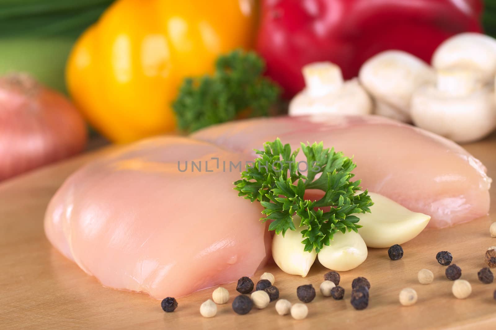 Raw chicken breast with white and black pepper corns, parsley leaf and garlic and with some fresh vegetable in the back (Selective Focus, Focus on the front of the meat and the parsley)