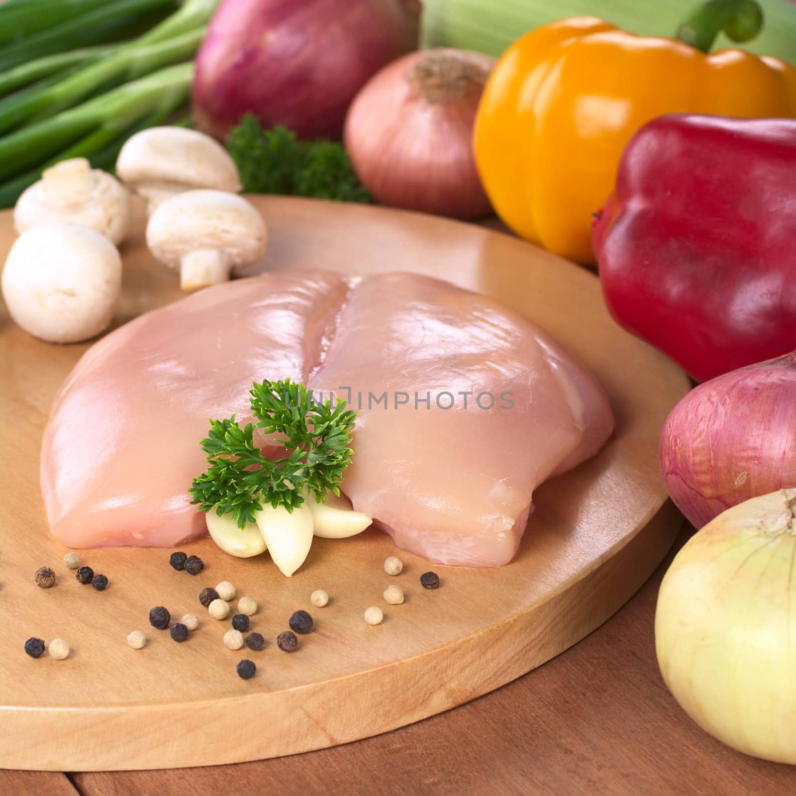 Raw chicken breast with white and black pepper corns, parsley leaf and garlic surrounded by fresh vegetable (Selective Focus, Focus on the front of the meat, the garlic and the parsley)