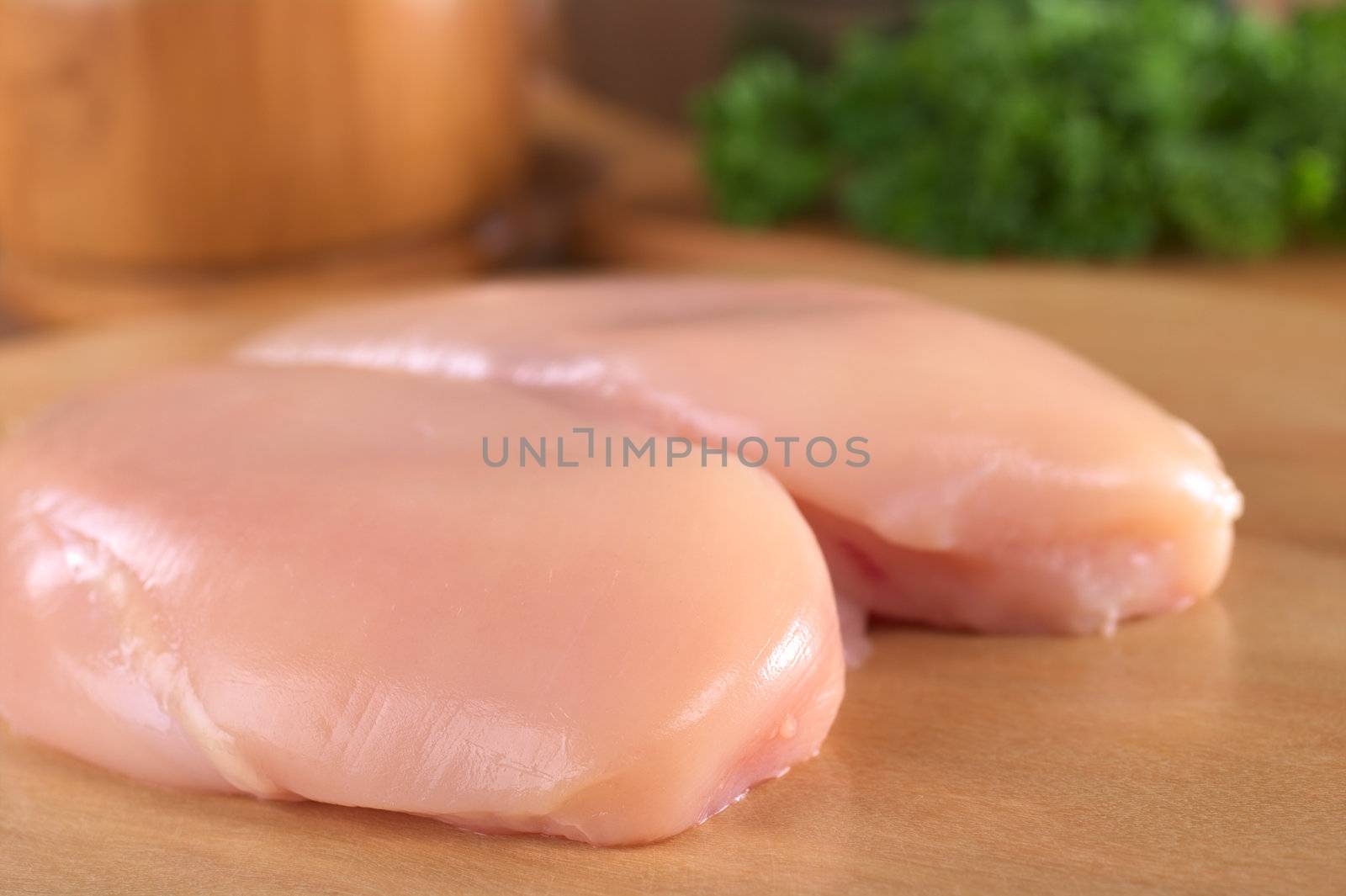 Raw chicken breast with wooden mortar and fresh parsley in the back (Selective Focus, Focus on the front of the meat)