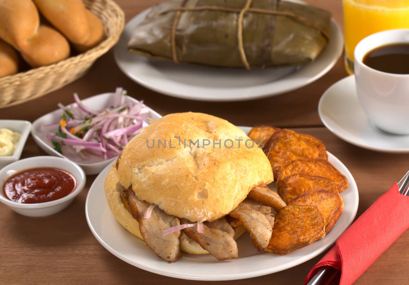 Typical Peruvian breakfast consisting of Pan con Chicharron (Bun with fried meat) and fried sweet potato, tamal (in the back), salsa criolla (onion salad) with coffee, orange juice and buns (Selective Focus, Focus on the front)