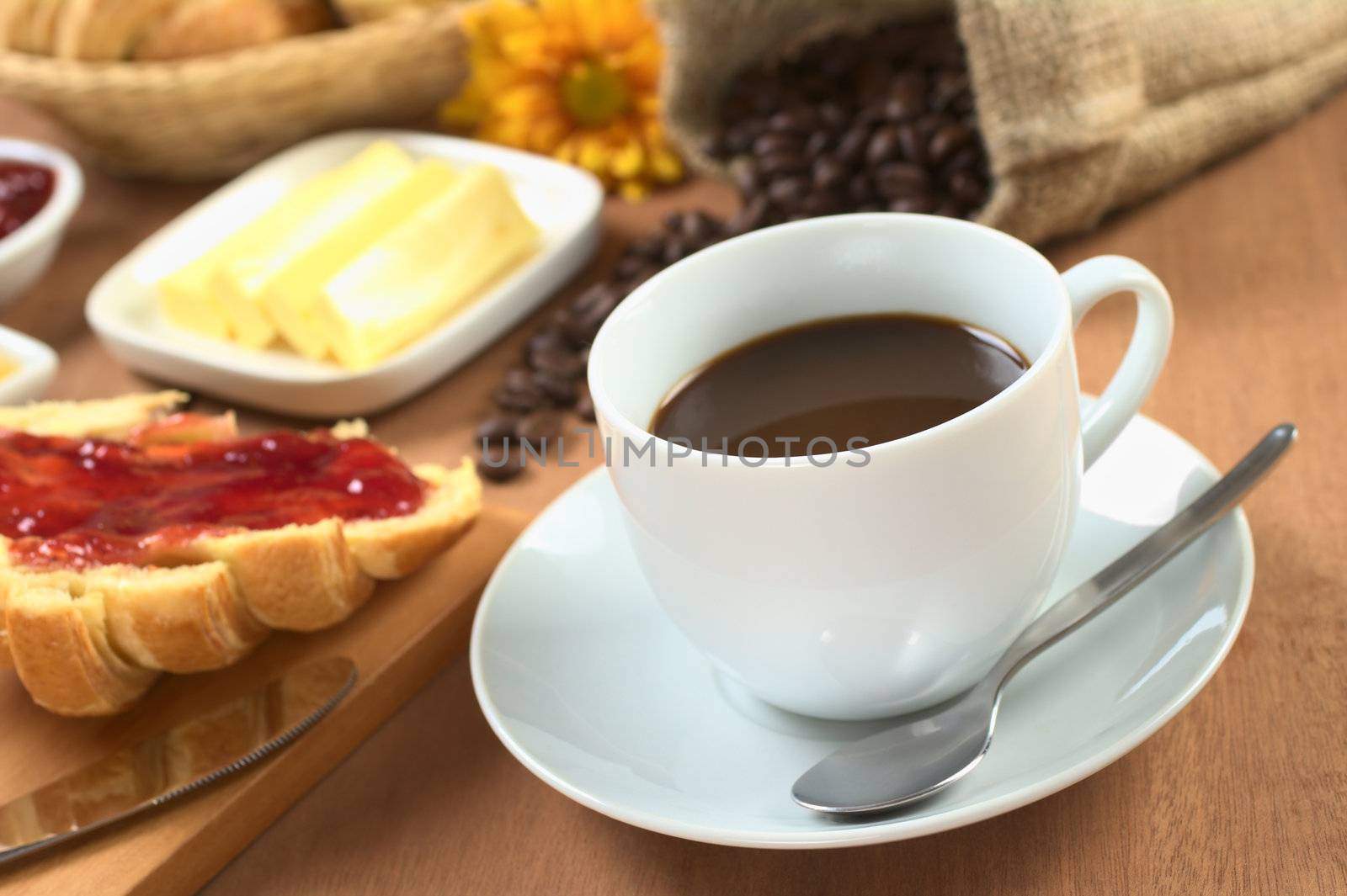 Hot, fresh coffee with croissant with strawberry jam, butter and coffee beans in the back (Selective Focus, Focus on the front rim of the coffee cup)