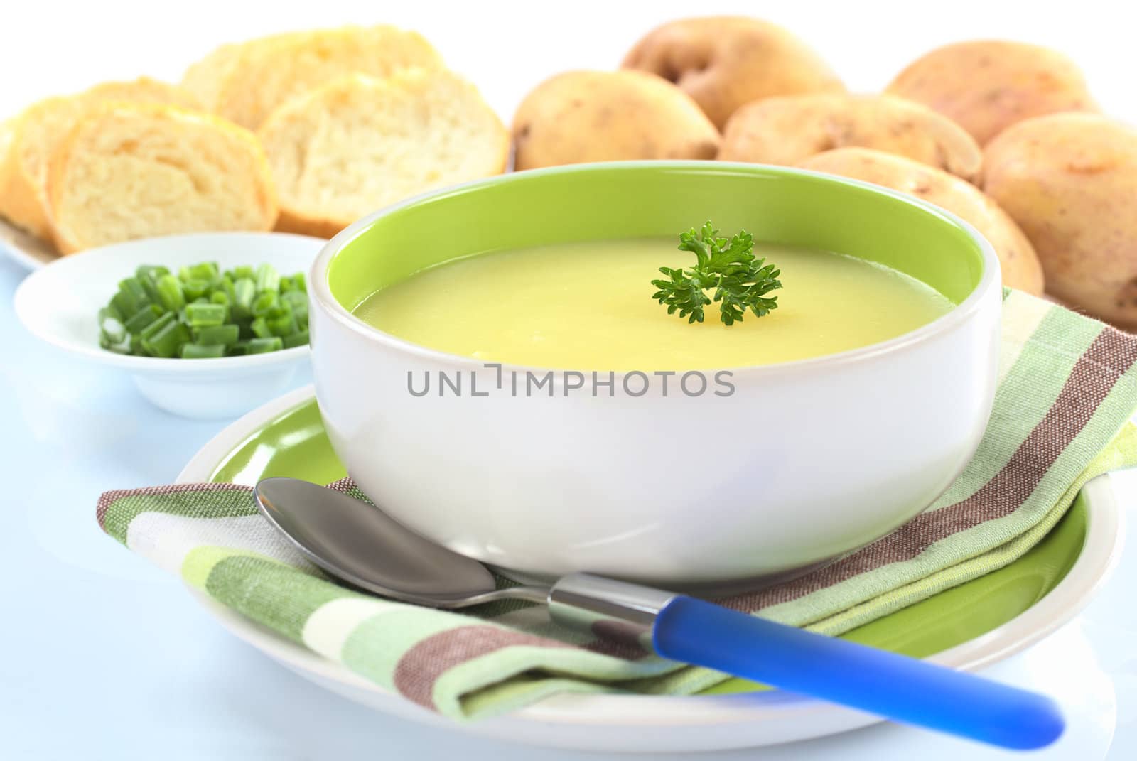 Fresh potato cream soup garnished with a parsley leaf with green onion, baguette slices and raw potatoes in the back on a slightly bluish surface (Selective Focus, Focus on the parsley leaf in the bowl) 