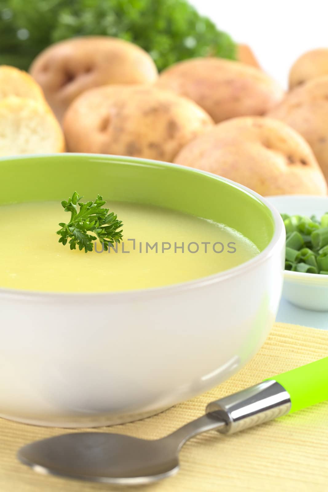 Fresh potato cream soup garnished with a parsley leaf with green onion, baguette slices, raw potatoes and parsley in the back (Selective Focus, Focus on the parsley leaf in the bowl) 
