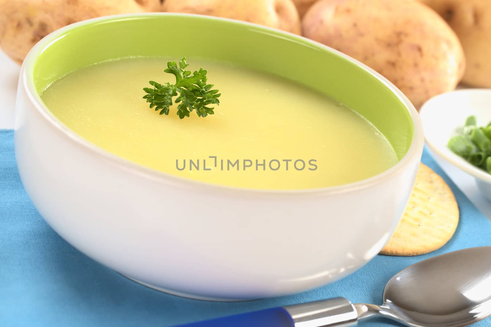 Fresh potato cream soup garnished with a parsley leaf with green onion and raw potatoes in the back on a blue table mat (Selective Focus, Focus on the parsley leaf in the bowl) 