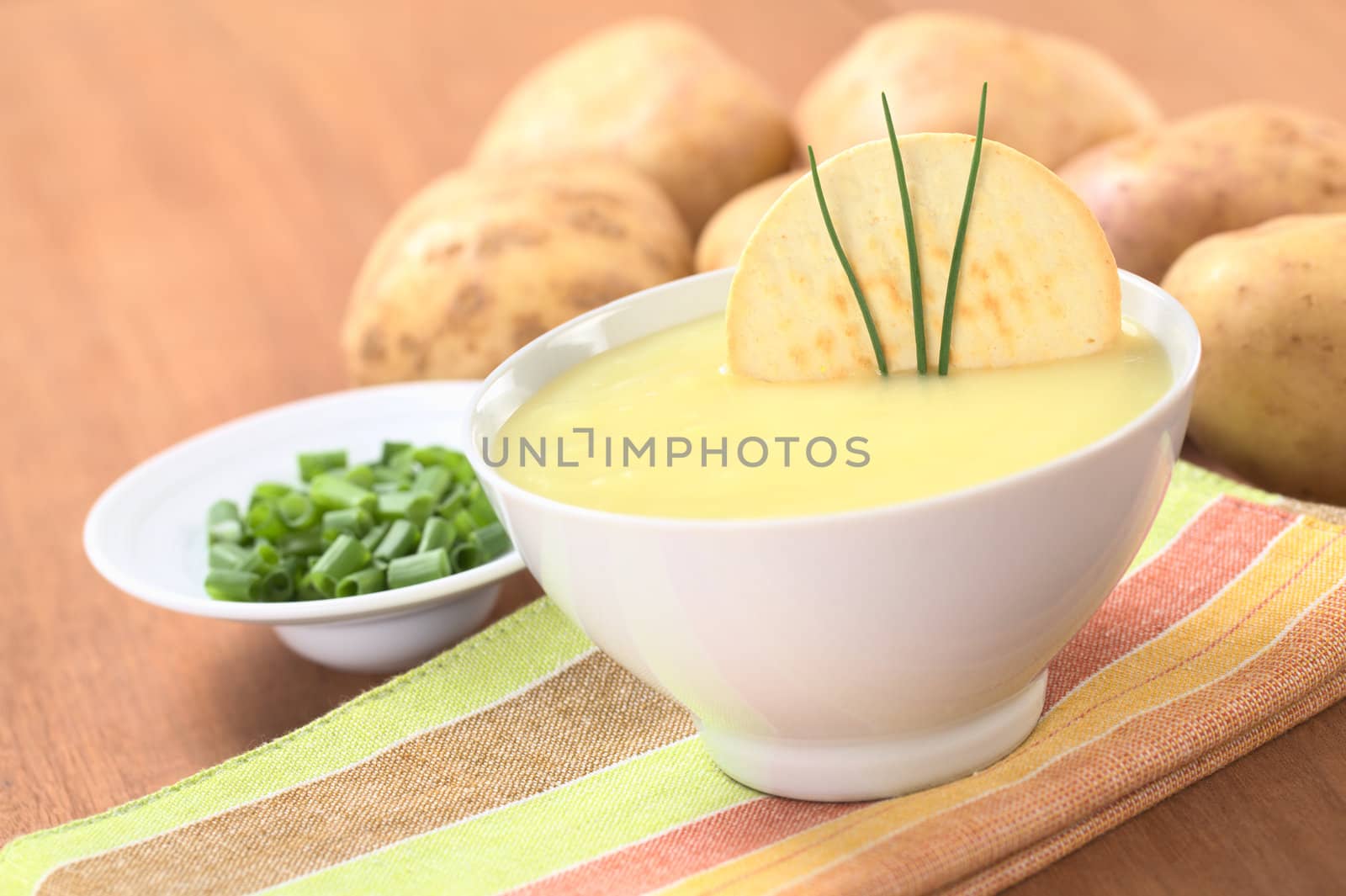 Fresh potato cream soup garnished with a cracker and chives with green onion and raw potatoes in the back on wood (Selective Focus, Focus on the cracker and the chives) 