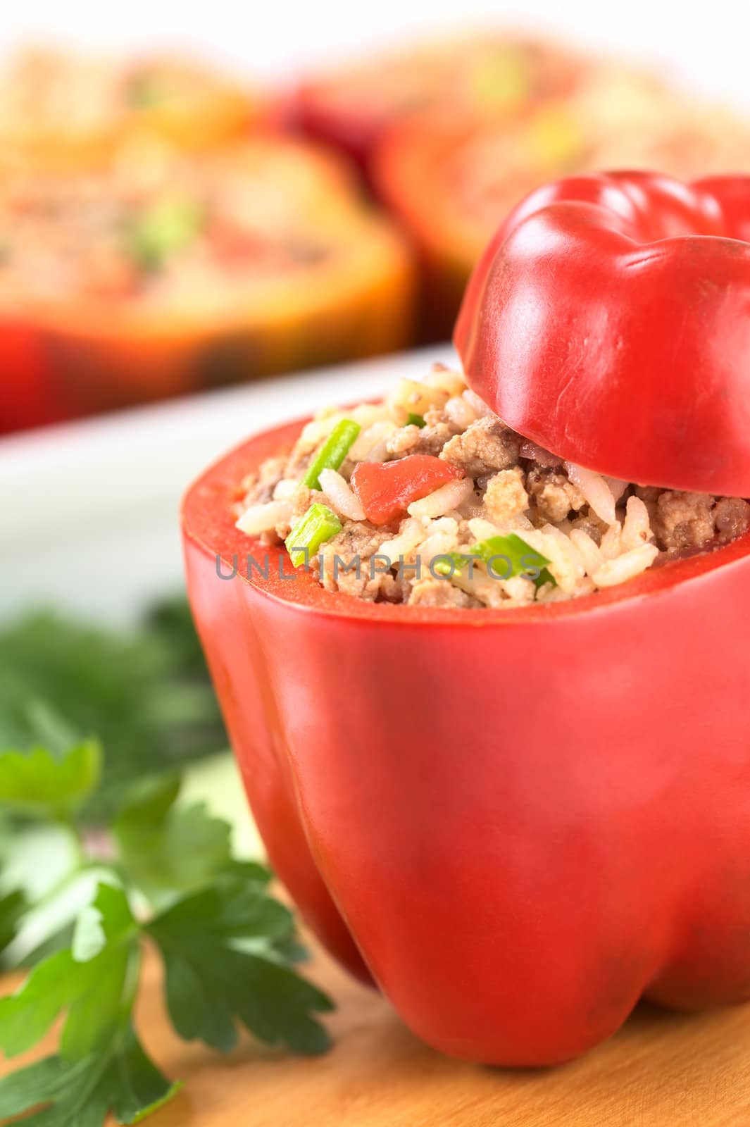 Unbaked stuffed red bell pepper filled with minced meat, onion, rice, tomato and green onion with parsley on the side and a casserole in the back (Selective Focus, Focus on the tomato piece and the stuffing around it in the pepper) 