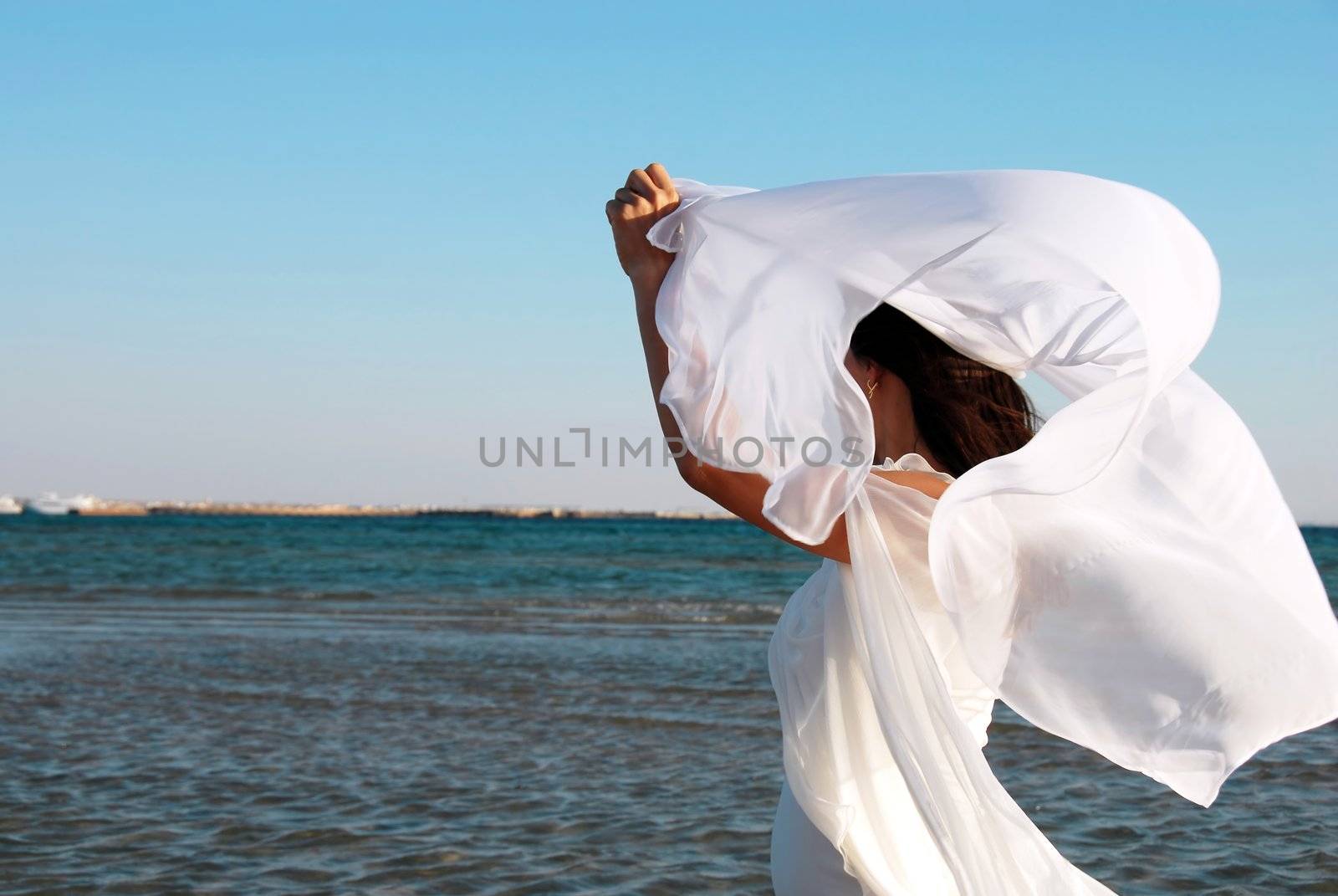 woman in white dress with flying scarf and raised hands on seaside