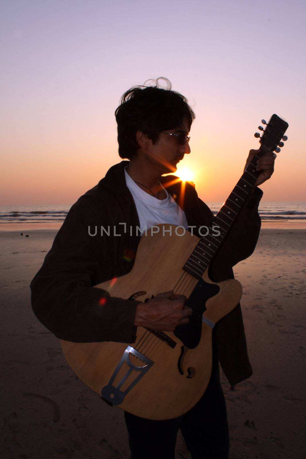 A young Indian guitarist performing on a beach at sunset.