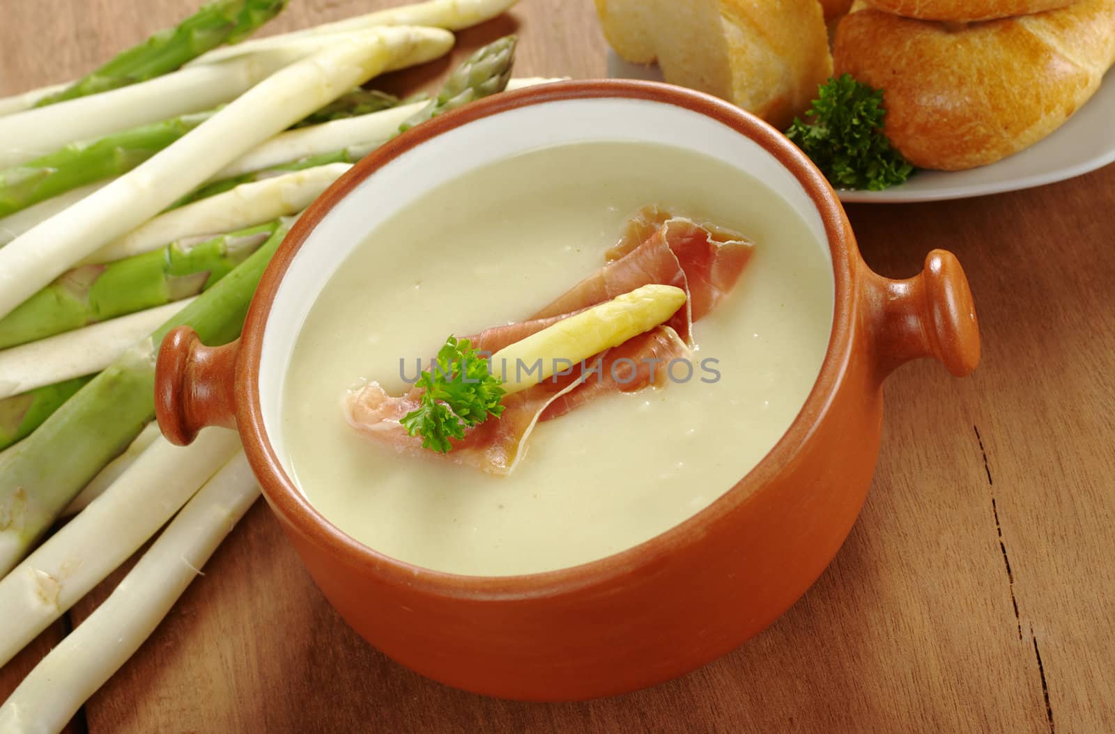 Asparagus soup garnished with ham, asparagus head and parsley accompanied by bread with raw asparagus lying by the side. All served in rustic style (Selective Focus, Focus on the garnish on the top of the soup) 
