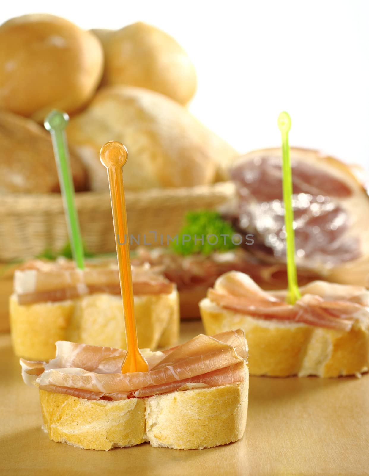 Baguette pieces with thin ham slices on top on plastic skewer with bread basket and big ham piece in the back (Selective Focus, Focus on front)