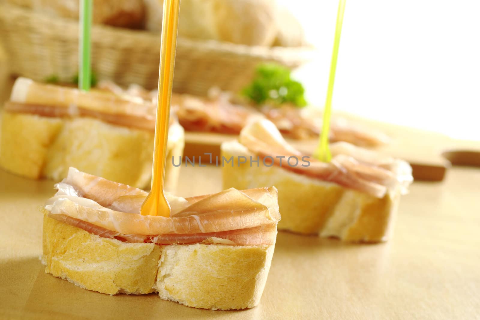 Baguette pieces with thin ham slices on top with plastic skewer and bread basket in the back (Selective Focus, Focus on front)