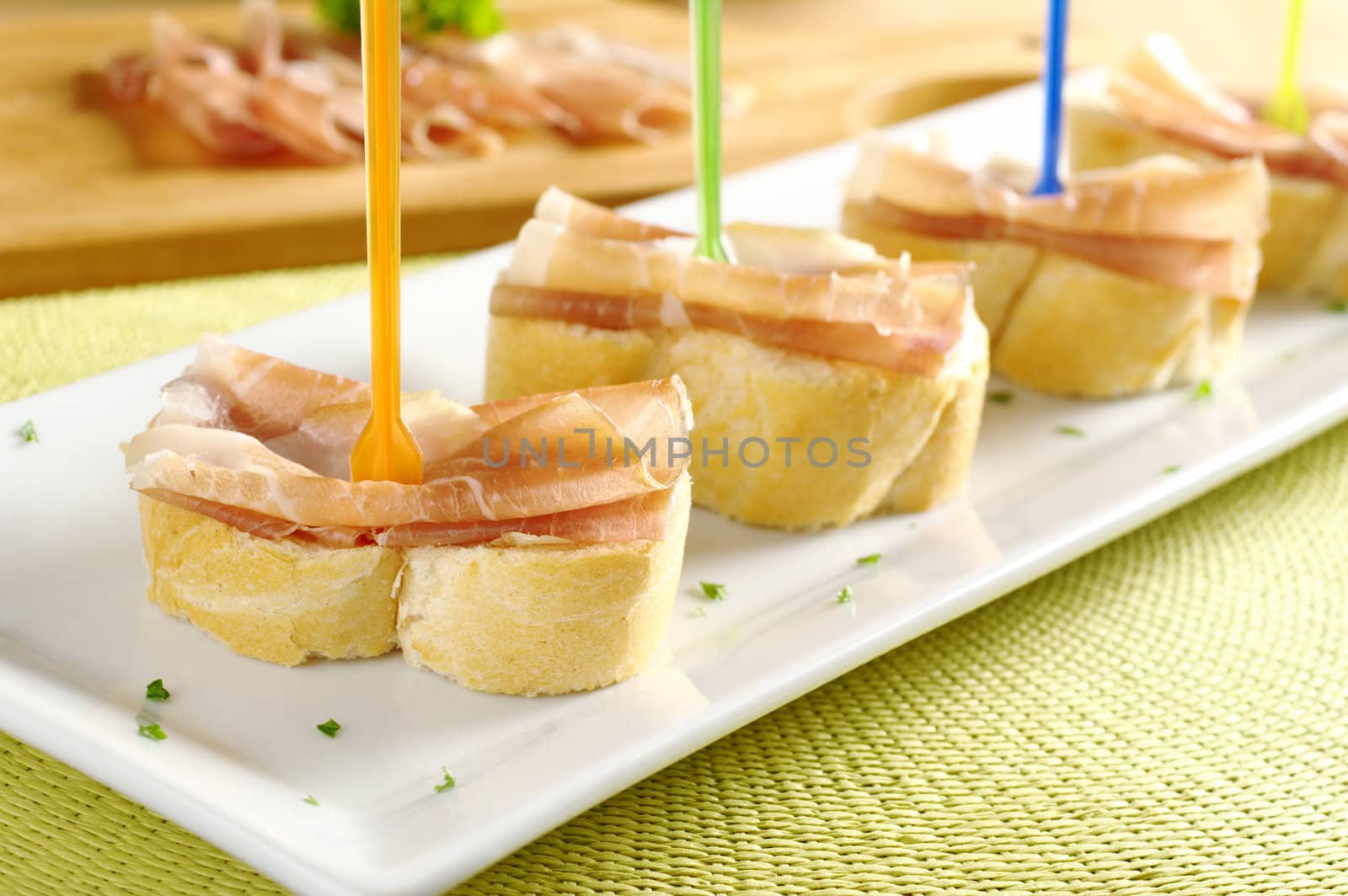 Baguette with thin ham slices on top with plastic skewer served on a long white plate with ham on cutting board in the back (Selective Focus, Focus on the front)