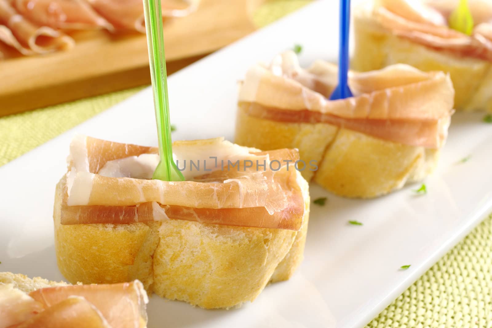 Baguette with thin ham slices on top with plastic skewer served on a long white plate with ham on cutting board in the back (Selective Focus, Focus on the ham with the green skewer)