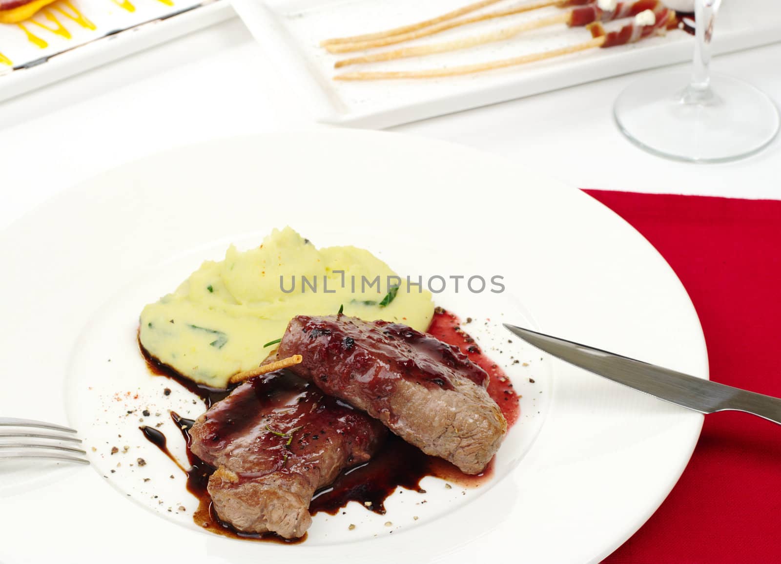 Main dish: Meat with red pepper sauce and mashed potato with cutlery as well as appetizers and wine glass in the background  (Selective Focus, Focus on the meat)