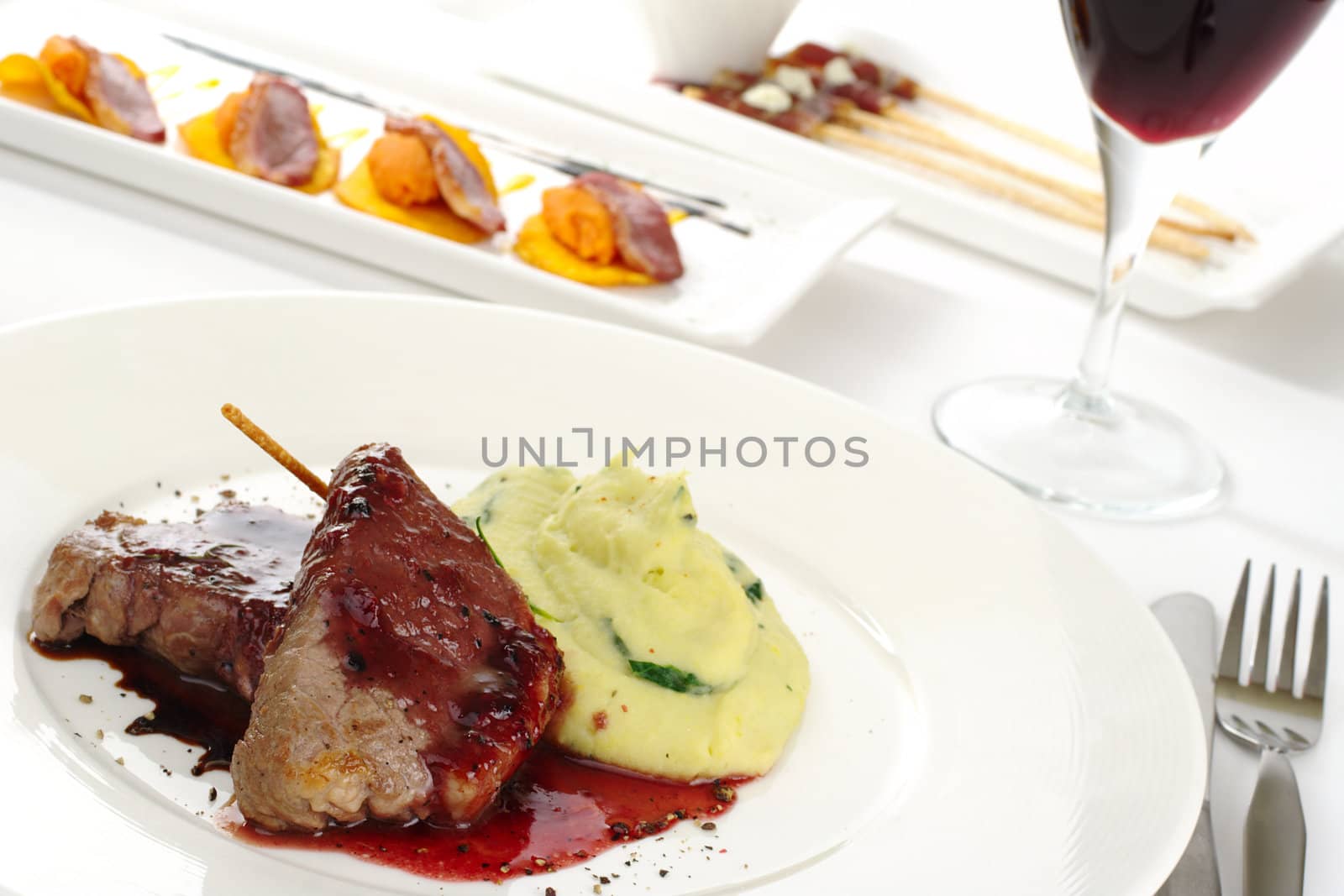 Main dish: Meat with red pepper sauce and mashed potato with cutlery as well as appetizers and red wine in the background  (Selective Focus, Focus on the meat)