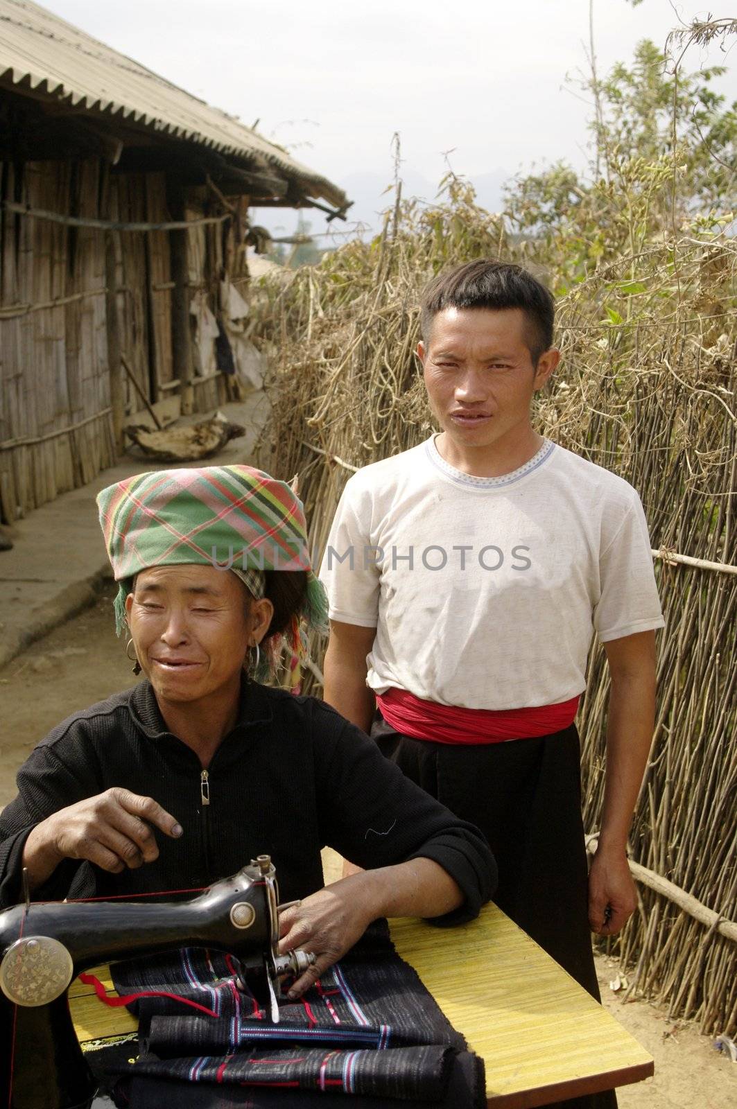 Black Hmong woman at her sewing machine. It is installed in front of his home. His hands are blue dyeing fabrics she sews. This machine is the sole property of the couple