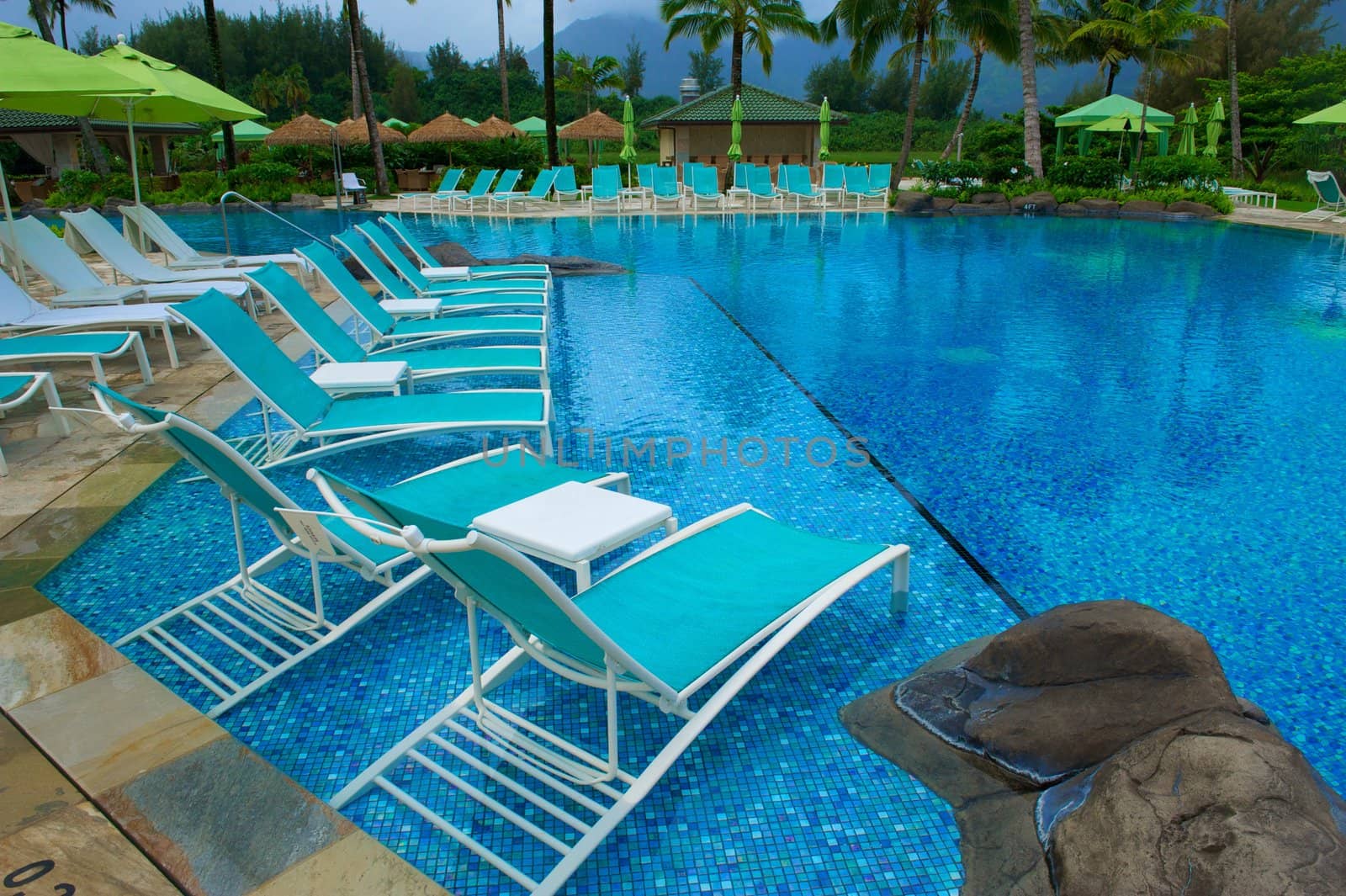 Turquoise chairs sitting in a deep blue swimming pool surrounded by green umbrellas at an upscale resort in Kauai, Hawaii