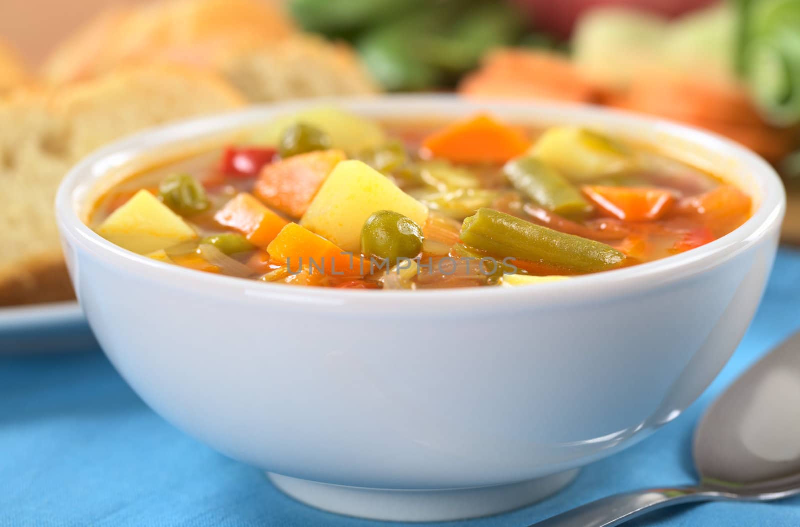 Fresh vegetable soup made of green bean, pea, carrot, potato, red bell pepper, tomato and leek in white bowl with baguette slices and ingredients in the back (Selective Focus, Focus on the vegetables one third into the soup)