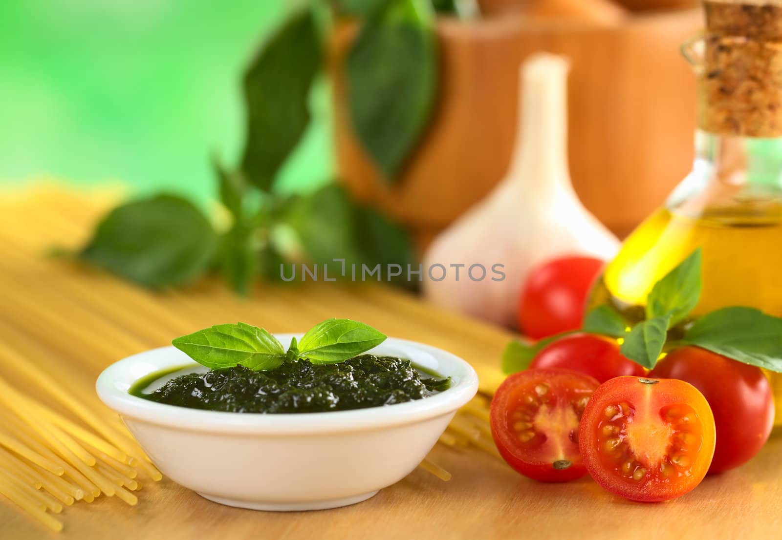 Fresh pesto made of basil and cherry tomatoes with olive oil, garlic, mortar, basil and raw spaghetti in the back (Selective Focus, Focus on the basil leaf on the pesto and the tomato in the front)