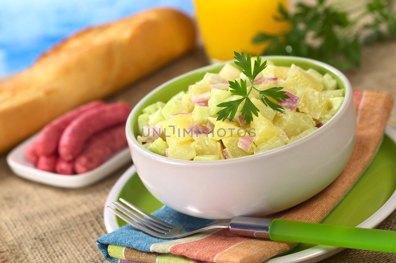 Potato salad made of cooked potatoes, red onions and cucumber, seasoned with a mayonnaise dressing and garnished with a parsley leaf with sausages, baguette and orange juice in the back (Selective Focus, Focus on the front of the salad and the leaf)