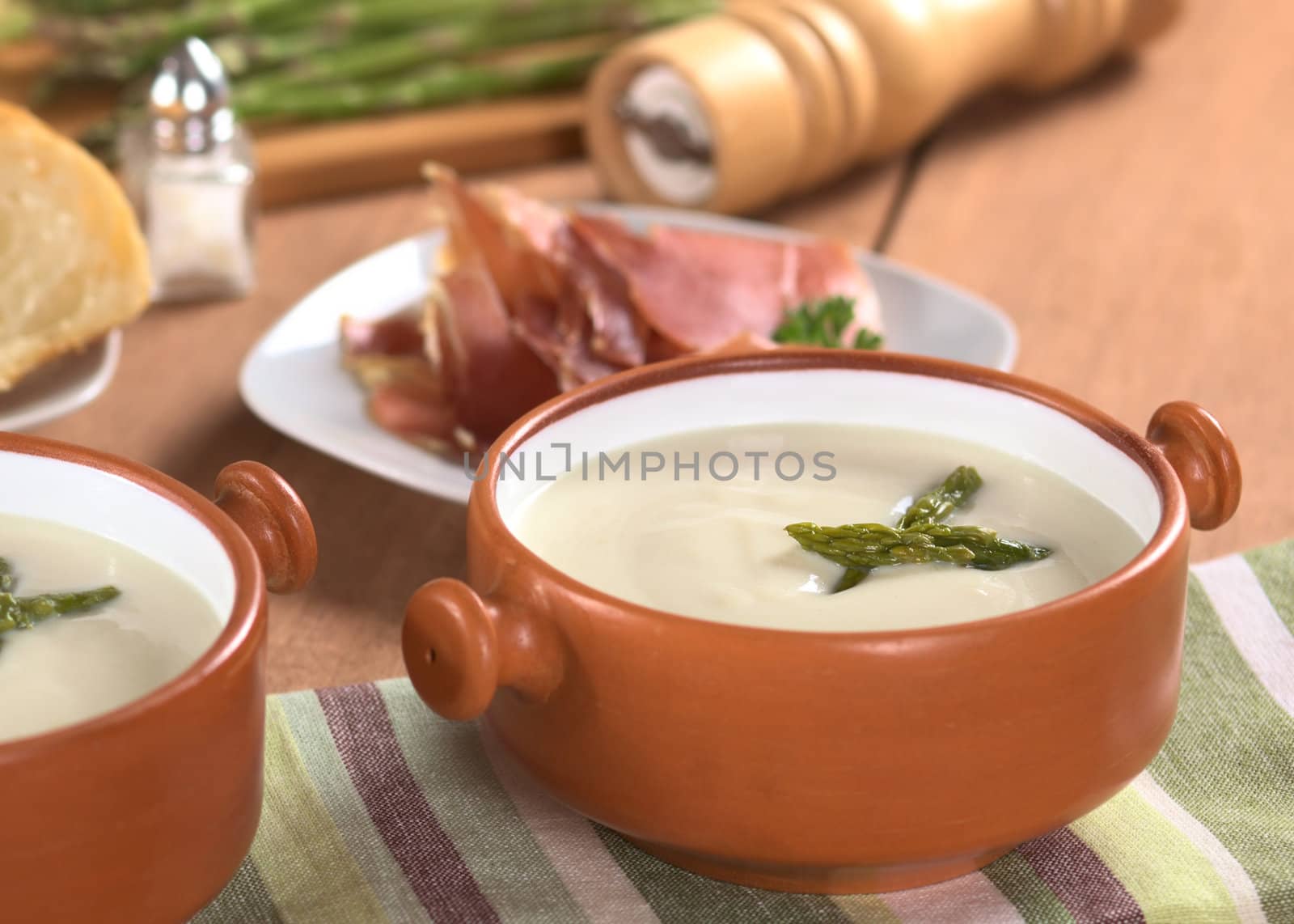 Cream of asparagus with green asparagus heads in rustic bowl with slices of ham, salt and pepper in the back on wood (Selective Focus, Focus on the asparagus head in the right bowl)
