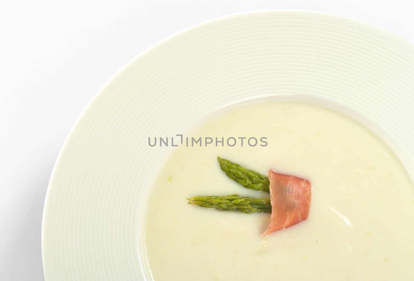 Cream of asparagus garnished with green asparagus heads and a slice of ham on top in a white soup plate (Selective Focus, Focus on the asparagus heads and the ham slice on the soup)