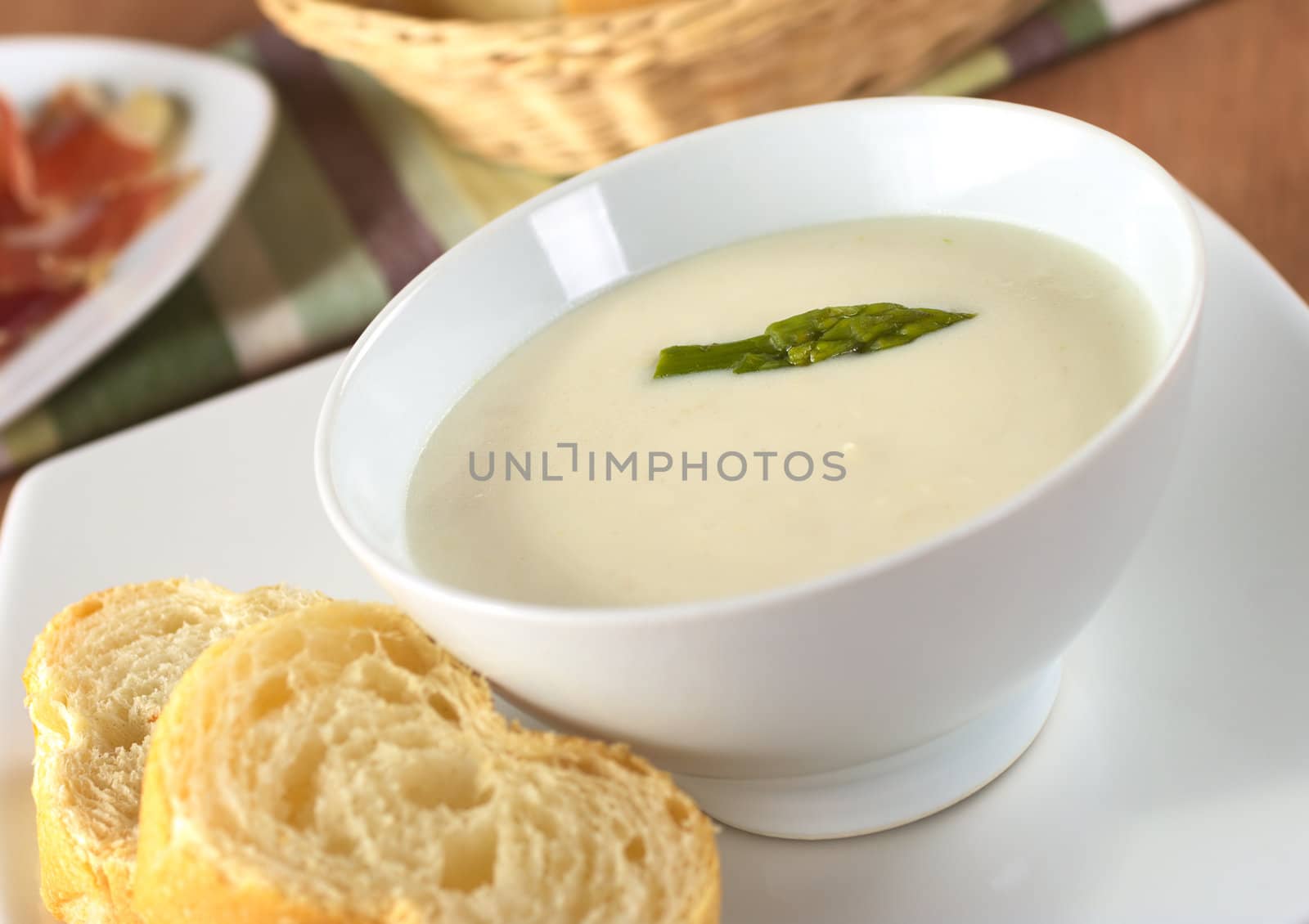 Cream of asparagus with a green asparagus head on top in a white bowl with baguette slices beside (Very Shallow Depth of Field, Focus on the asparagus head in the soup)