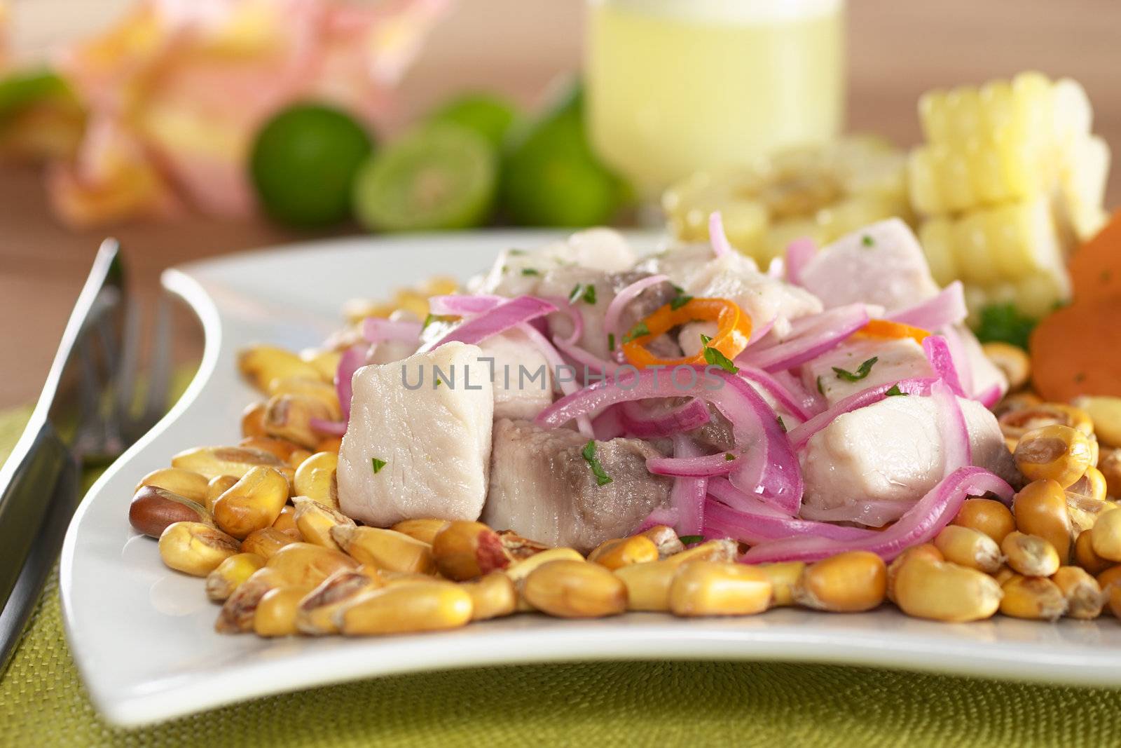 Peruvian-style ceviche made out of raw mahi-mahi fish (Spanish: perico), red onions and aji (Peruvian hot pepper) and served with roasted corn (cancha) and cooked corn. In the back Peruvian cocktail called "Pisco Sour", limes and a flower. (Selective Focus, Focus on the front of the fish)