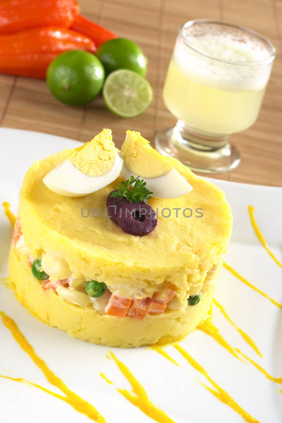 Traditional Peruvian dish called Causa made of mashed yellow and white potato mixed with aji (hot pepper) and lime juice and filled with vegetables (corn, peas, carrots) and mayonnaise and garnished with eggs and olives, with Pisco Sour in the back (Selective Focus, Focus on the front) 