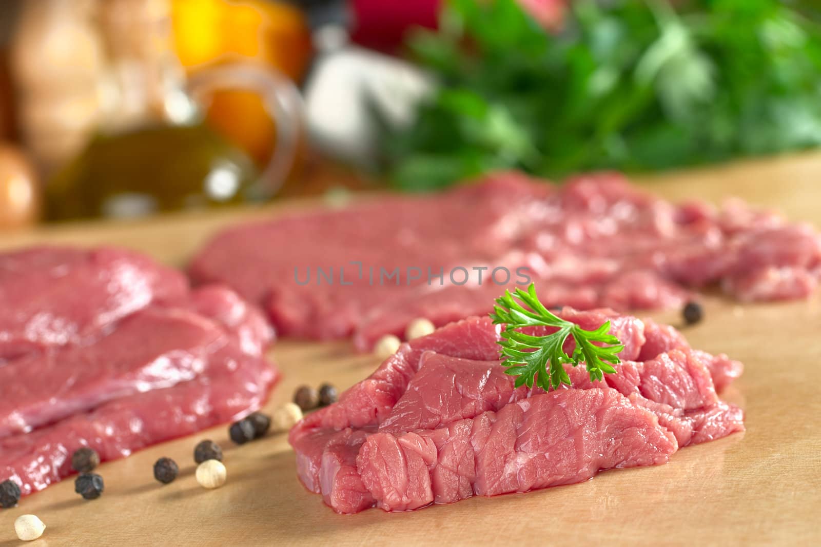 Fresh raw beef meat with parsley on top, with vegetables, herbs and kitchen utensils in the back (Selective Focus, Focus on the front of the meat and the parsley)