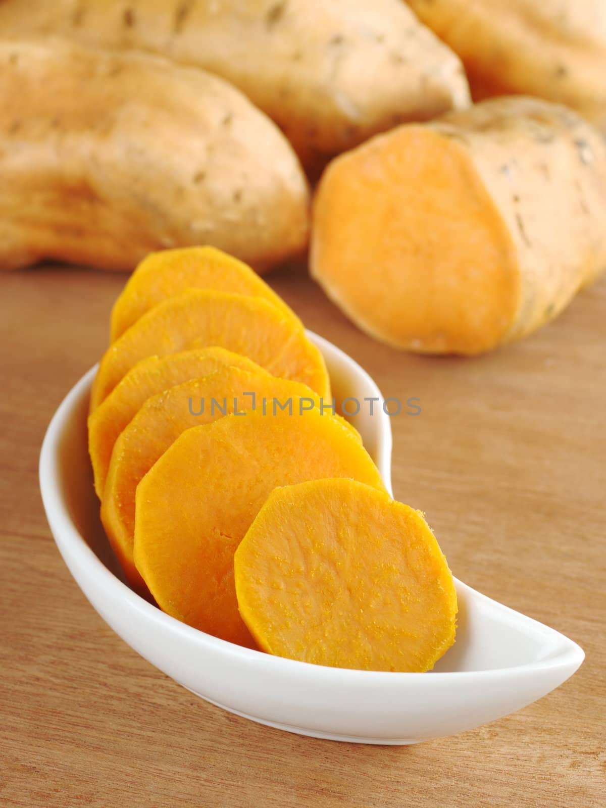 Cooked sweet potato (lat. Ipomoea batatas) cut in slices in white bowl on wooden surface with sweet potatoes in the background (Selective Focus, Focus on the front of the bowl) 