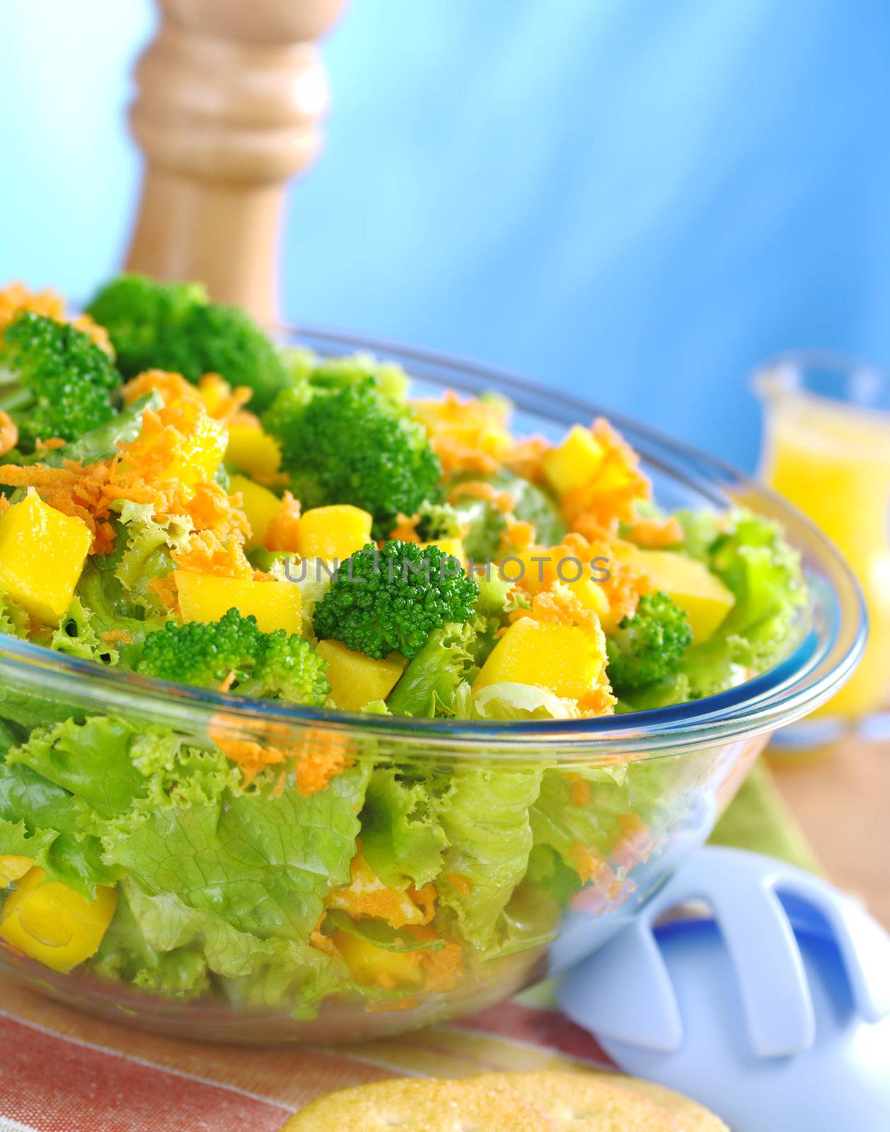 Broccoli-mango-carrot-lettuce salad in a glass bowl with salad servers laying next to it and a pepper mill and orange juice dressing in the background (Selective Focus, Focus on the front)