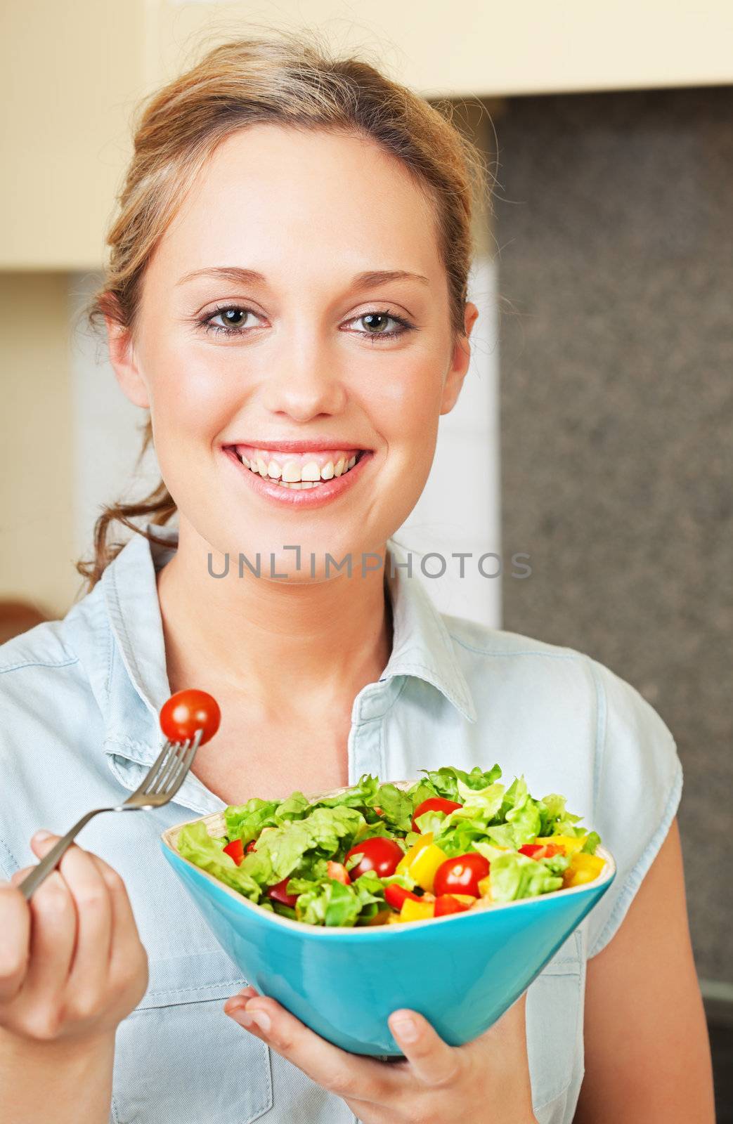 Pretty smiling young woman standing in the kitchen and holding a bowl with salad