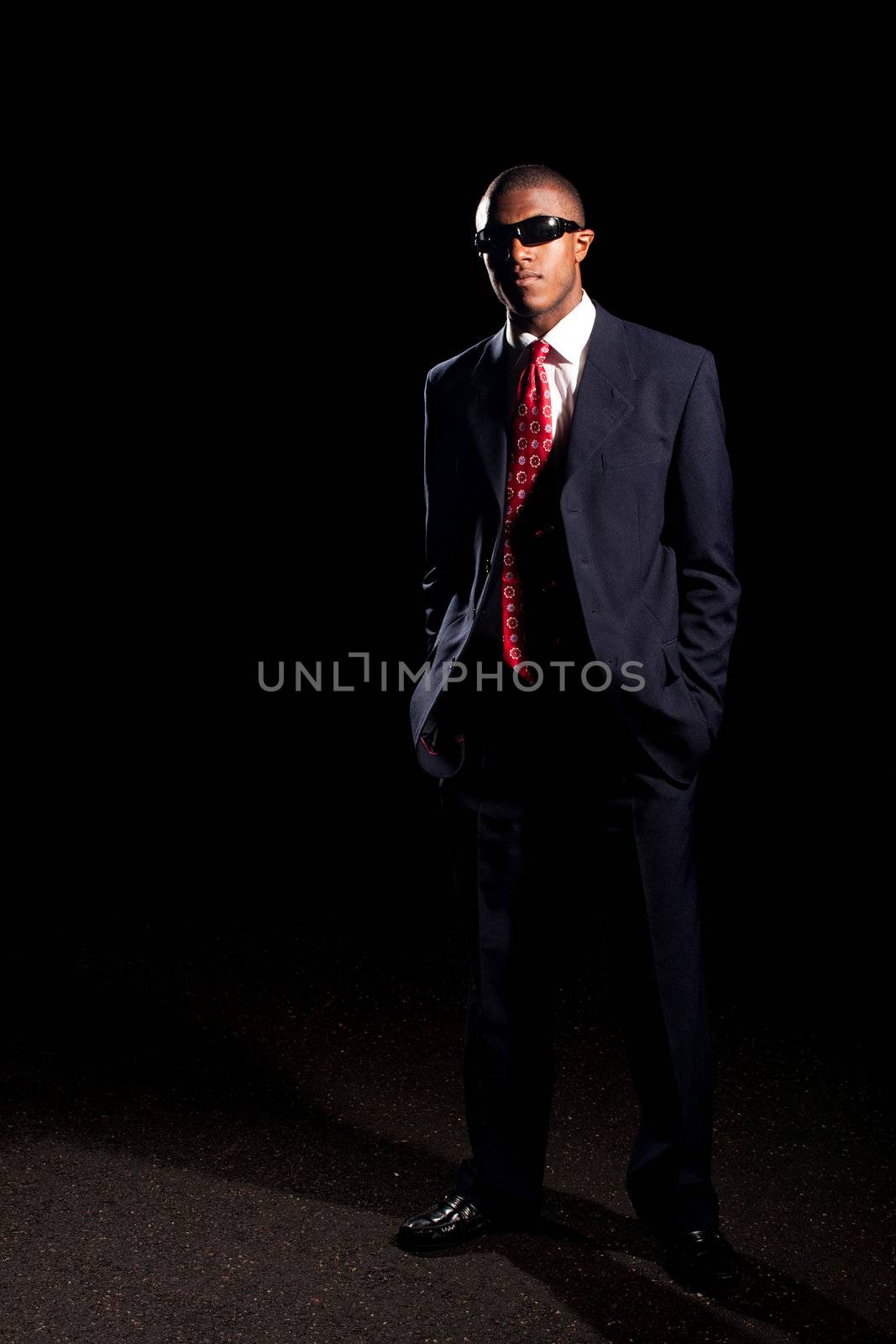  An African American man dressed in a dark colored suit and sunglasses standing in front of a dark black background.