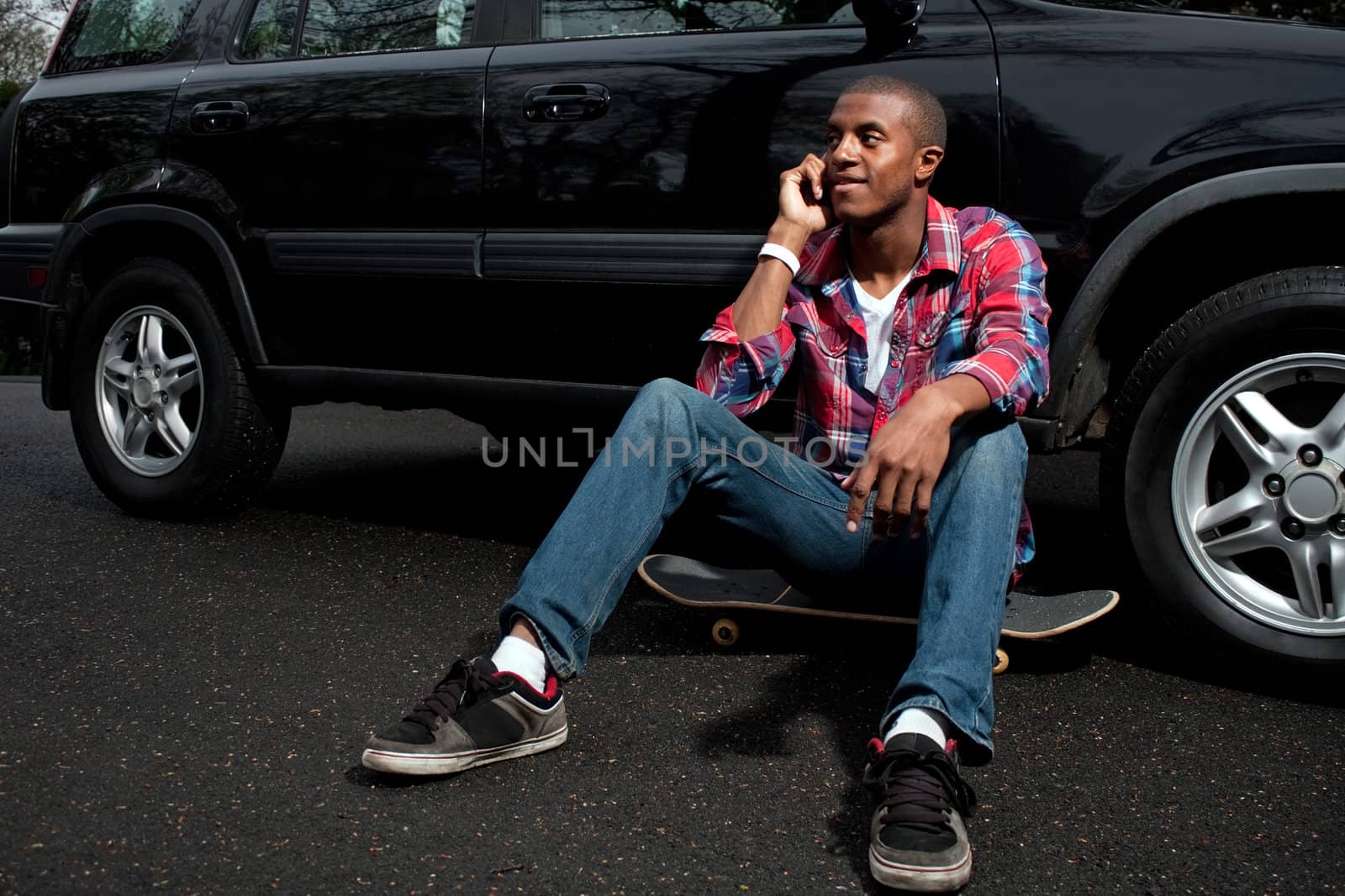 A young man hanging out sitting on his skateboard near his vehicle as he talks on his smartphone.